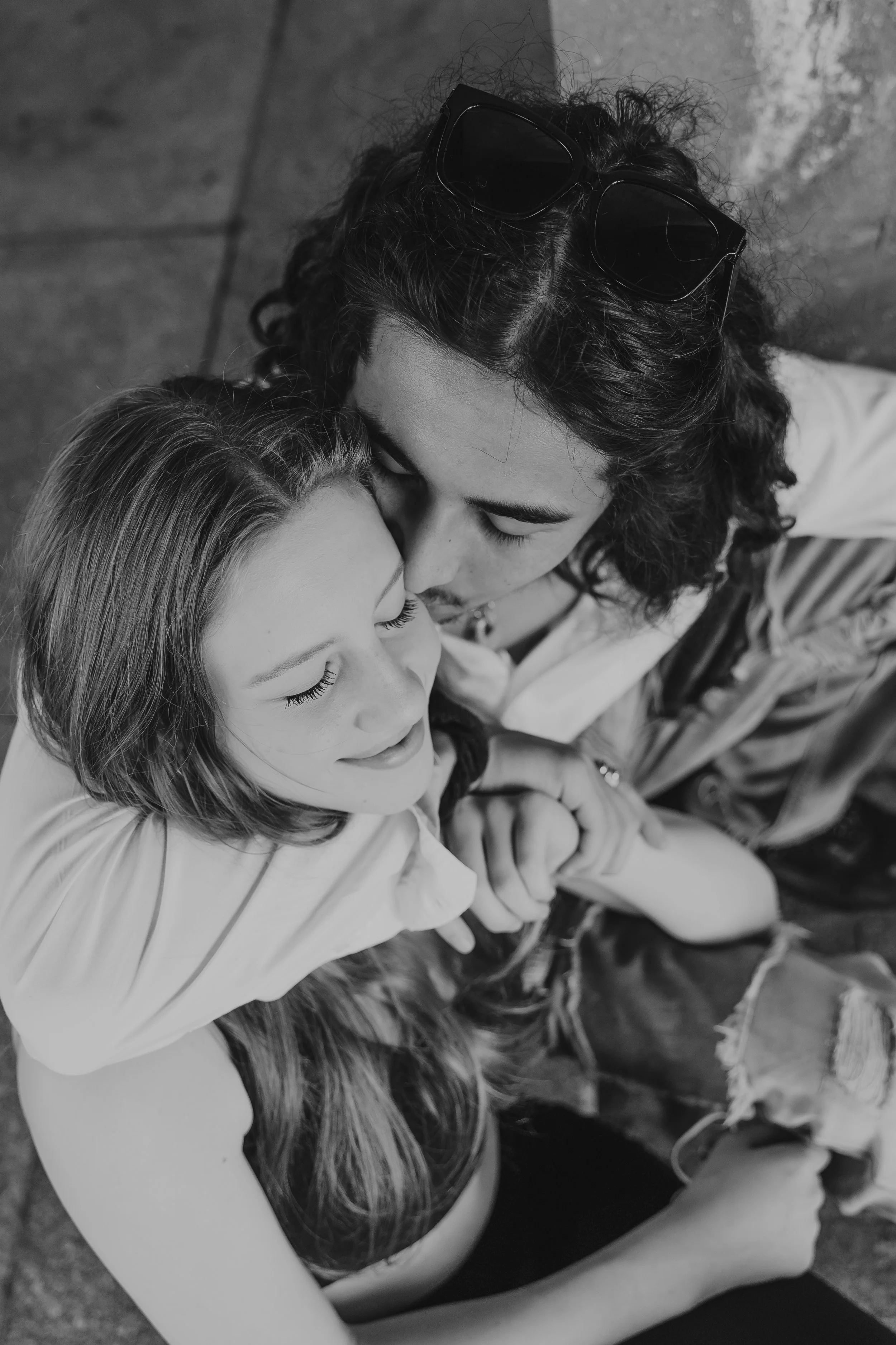Black and white photo of a couple sitting on the floor, the woman resting her head on the man's shoulder with eyes closed, the man leaning in with eyes closed, both smiling gently.