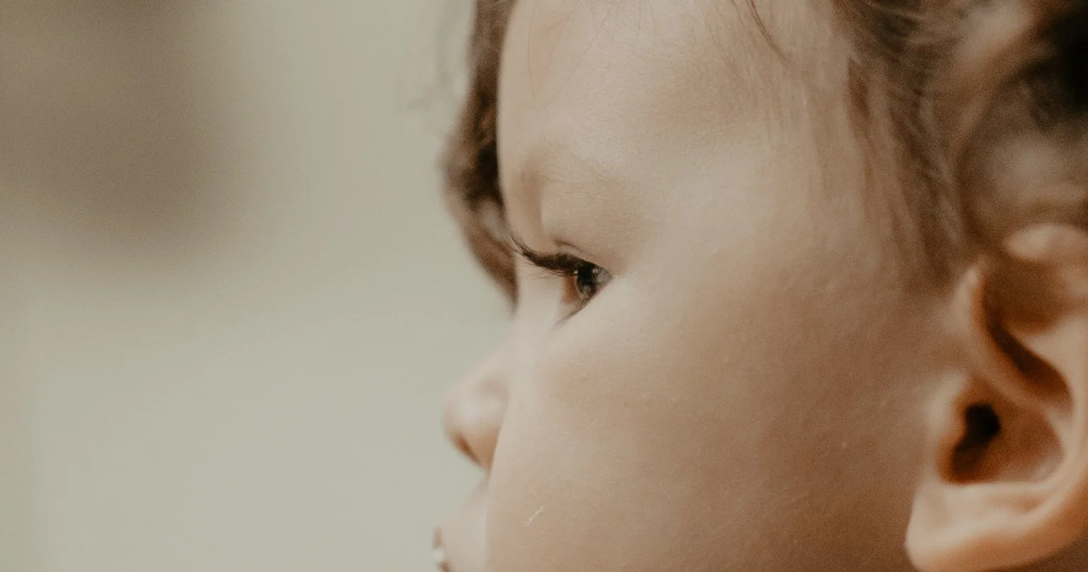 Close-up side profile of a young child's face showing an eye, cheek, and ear, with soft lighting and neutral background.
