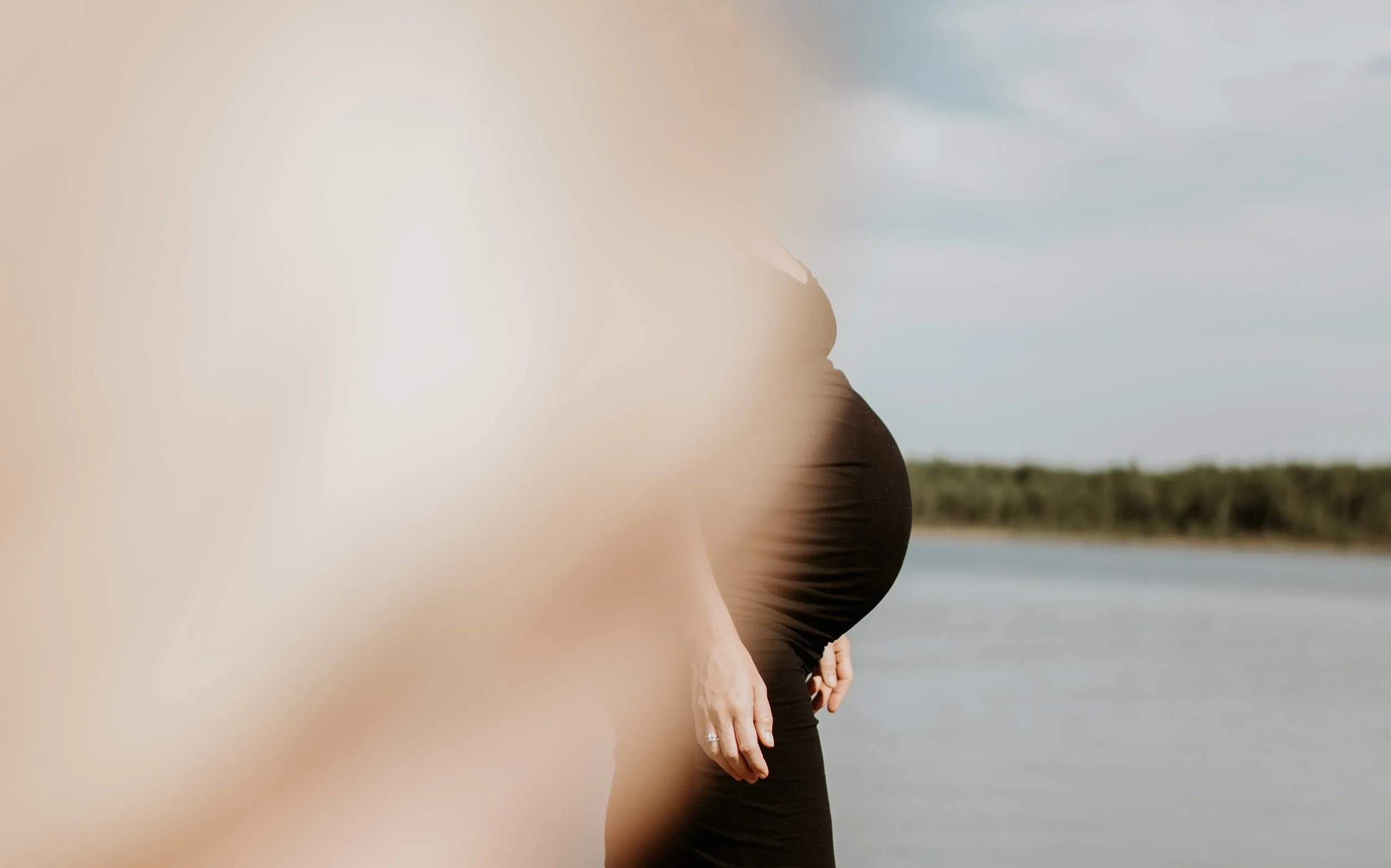 A pregnant woman in a black dress standing beside a body of water with trees in the background, partially obscured by a blurred foreground.