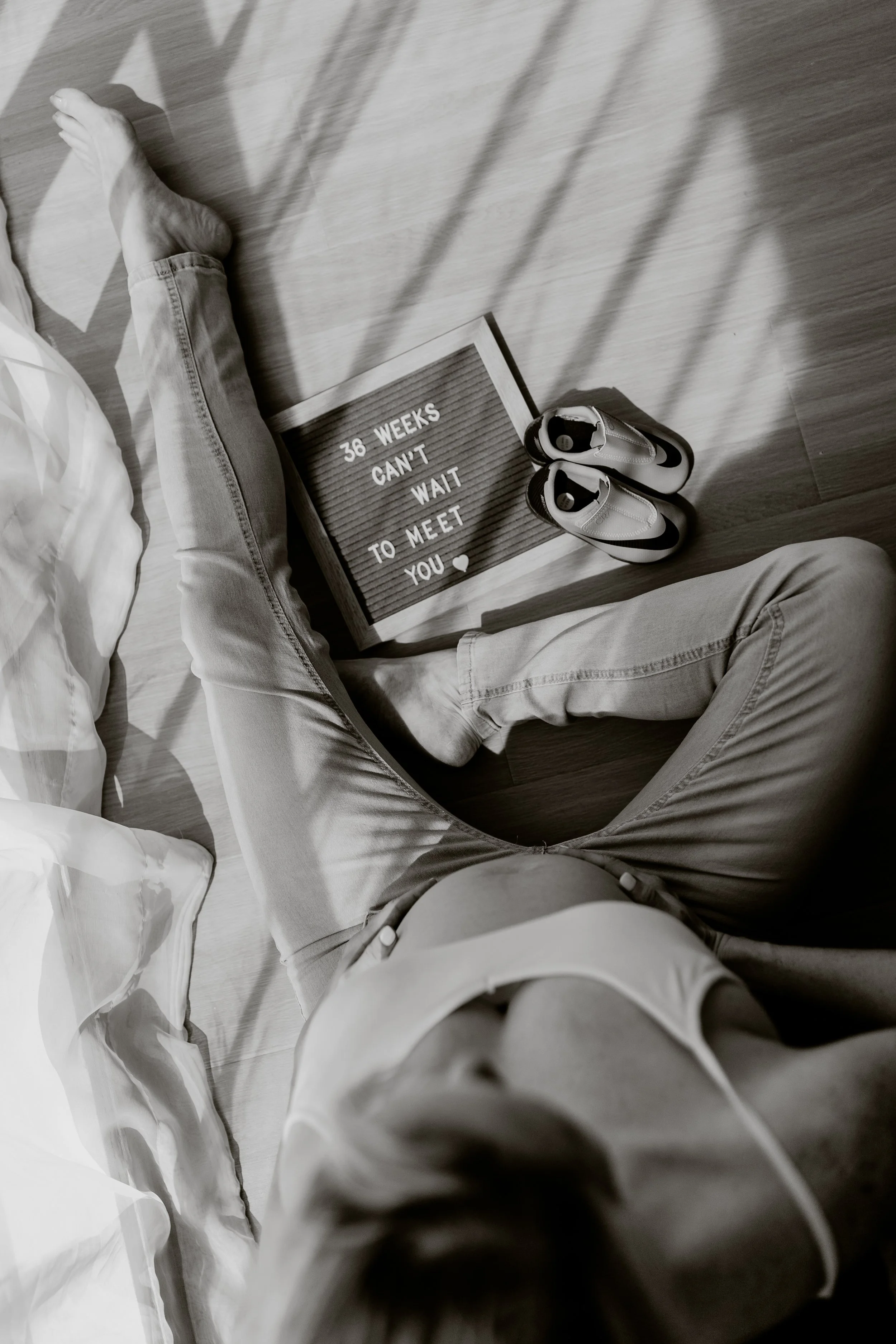 A person sitting cross-legged on a wooden floor, with a pair of sneakers nearby, a light box with the message "36 WEEKS CAN'T WAIT TO MEET YOU" and a small heart symbol.