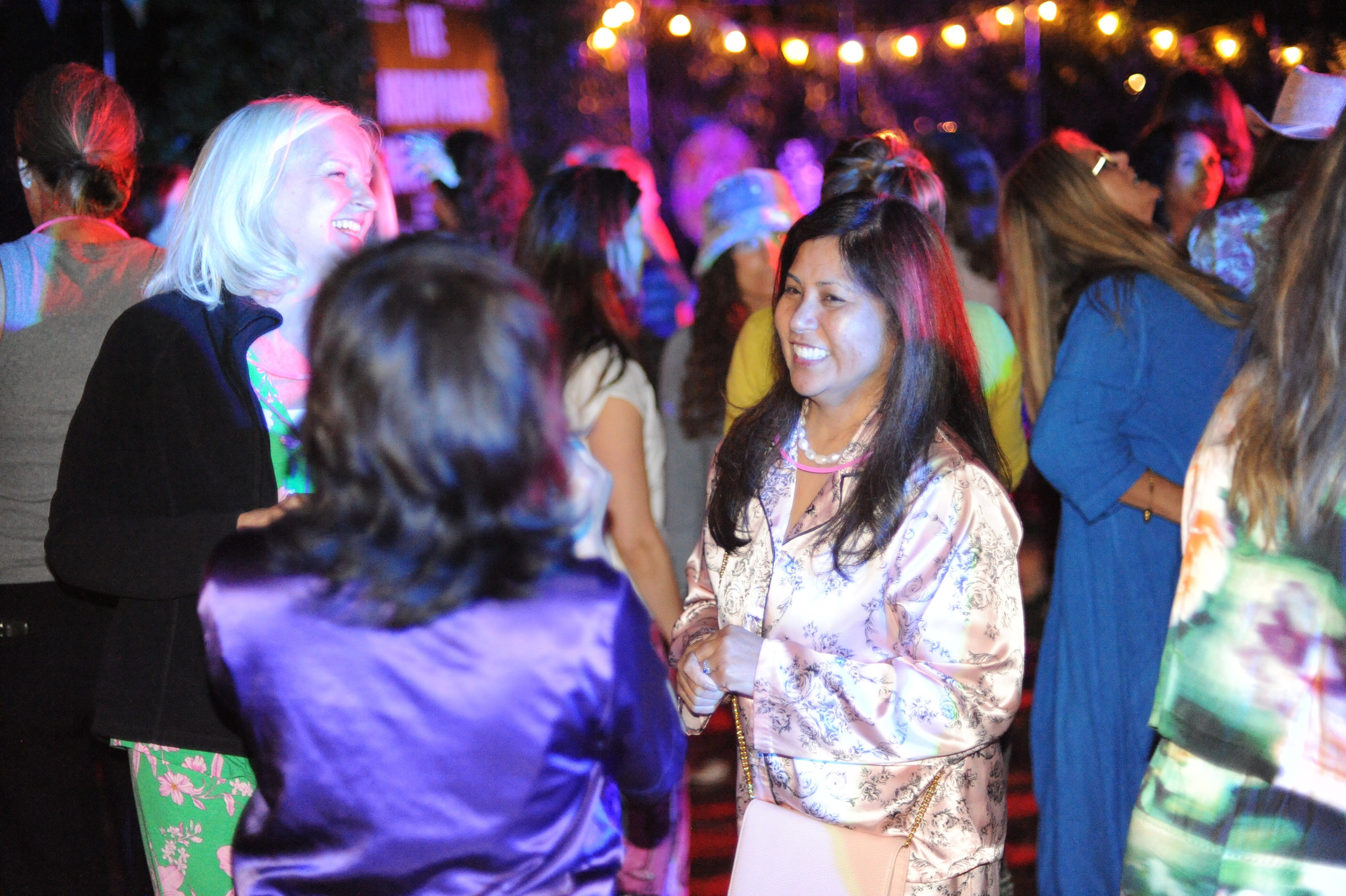 Group of women socializing at a lively event with colorful lighting and string lights in the background.