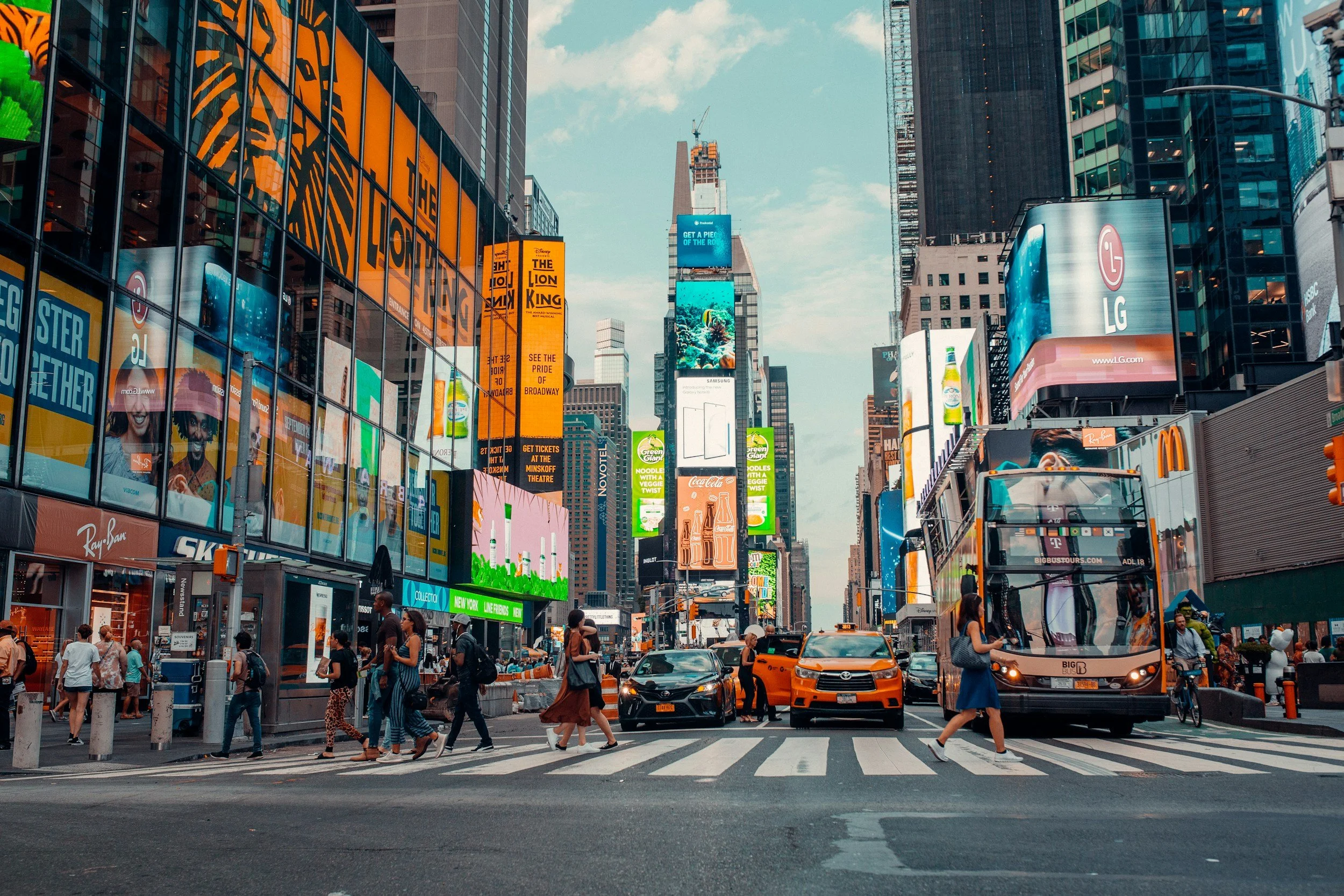 Busy city street in Times Square, New York City, with pedestrians crossing the street, yellow taxis, a double-decker bus, and many digital billboards displaying advertisements, including for LG, Coca-Cola, and other brands.