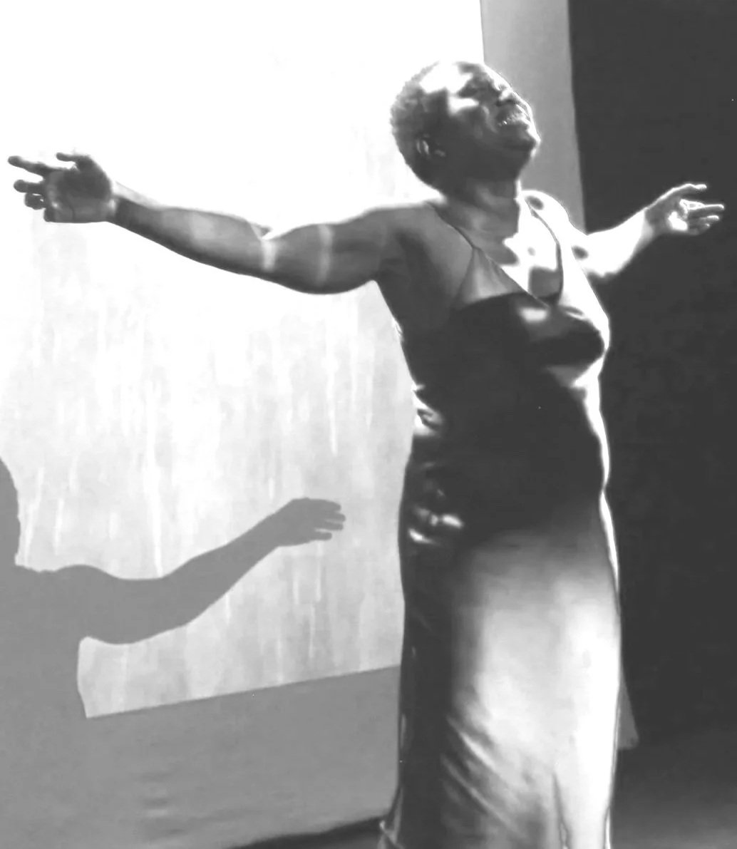 A woman in a red dress performing on stage with her arms outstretched and her shadow cast on the screen behind her.