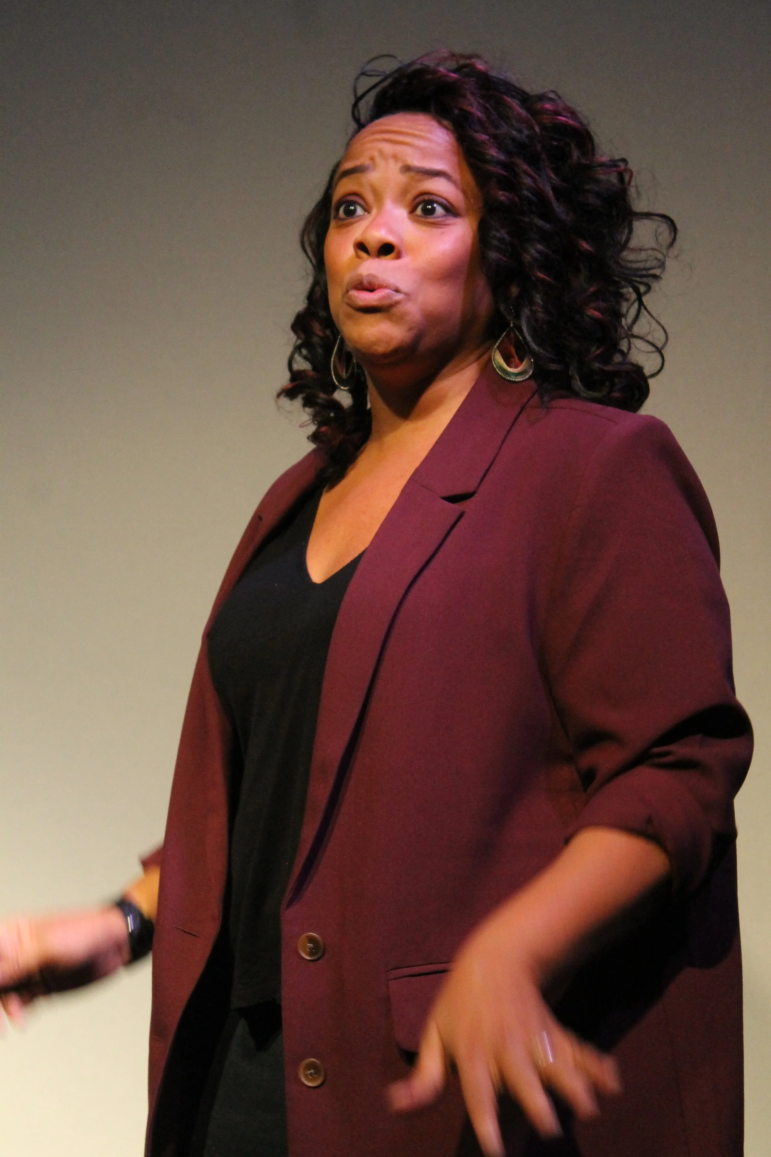 A woman with curly dark hair wearing a maroon blazer and black top, making a surprised or humorous facial expression.