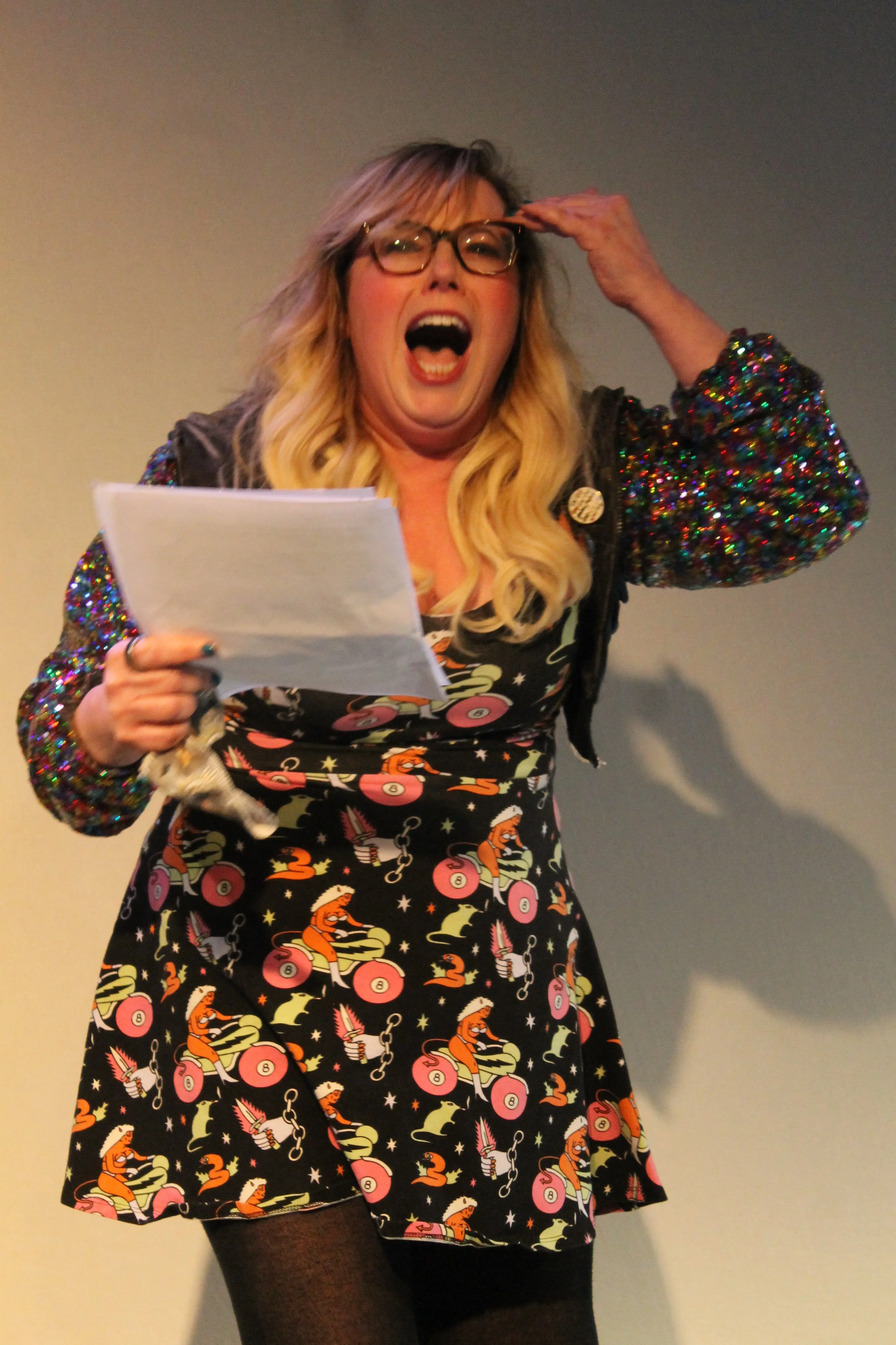 A woman with blonde hair and glasses, wearing a colorful sequin jacket and a dress with a cartoon pattern, appears to be speaking or protesting, holding papers in one hand and touching her forehead with the other.
