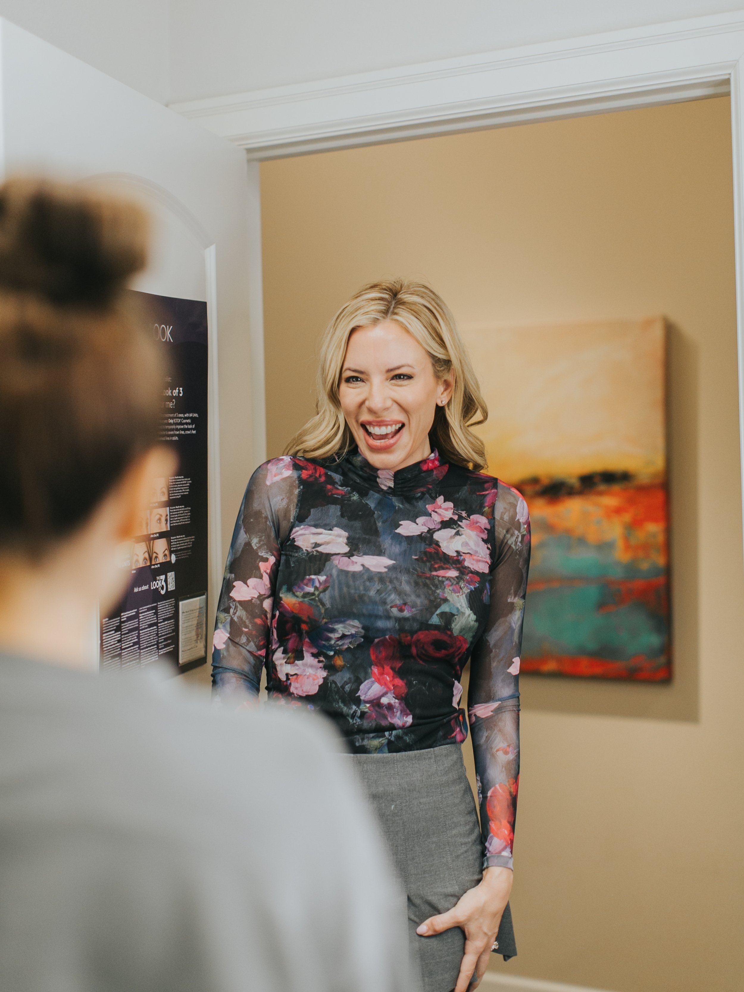 A woman in a floral shirt smiling in a well-lit room with a painting on the wall.