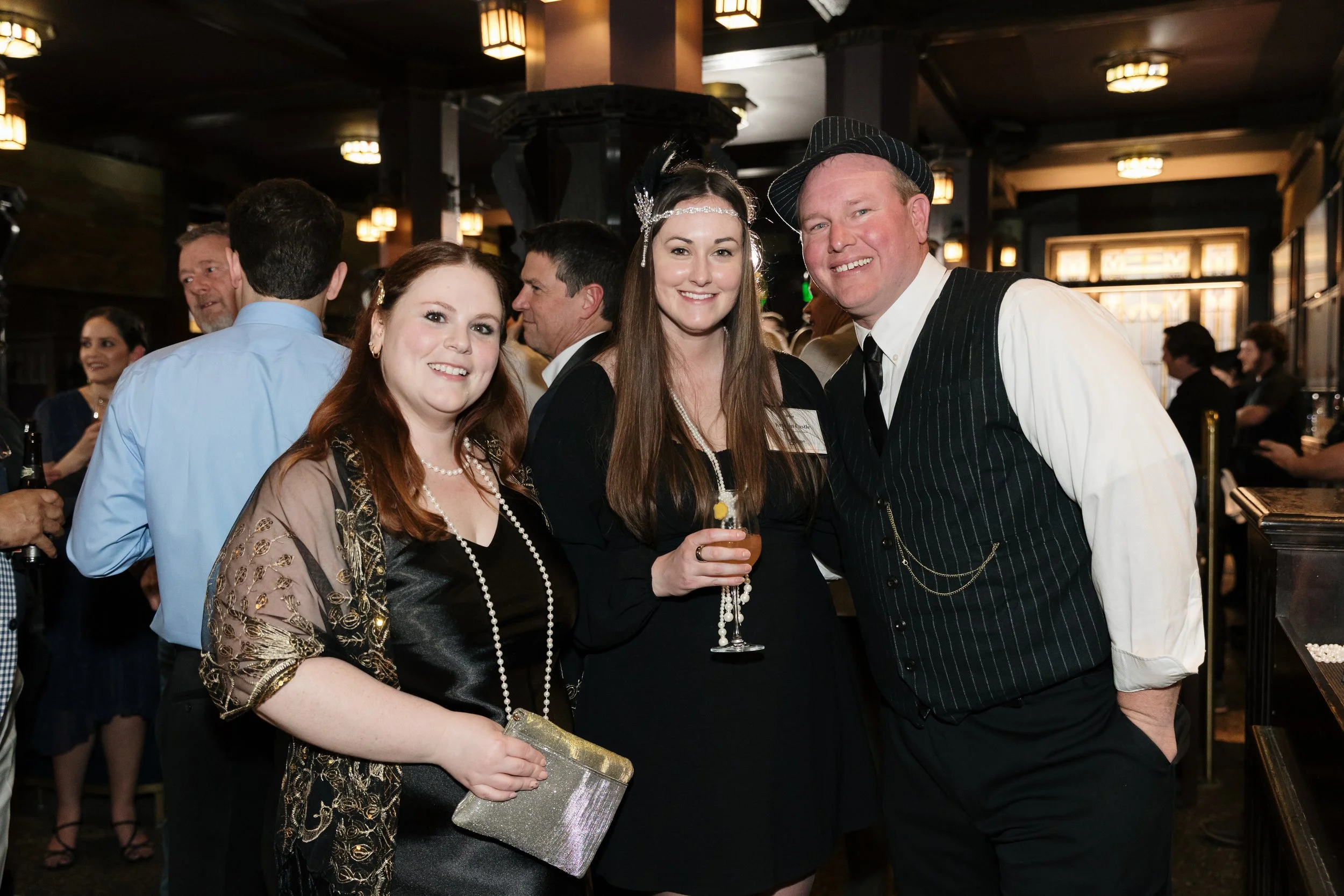 Group of people at a social event, including a woman with long brown hair holding a glass, a woman with long reddish-brown hair wearing a black dress and pearl jewelry, and a man with light-colored hair wearing a vintage-style pinstripe vest and hat,