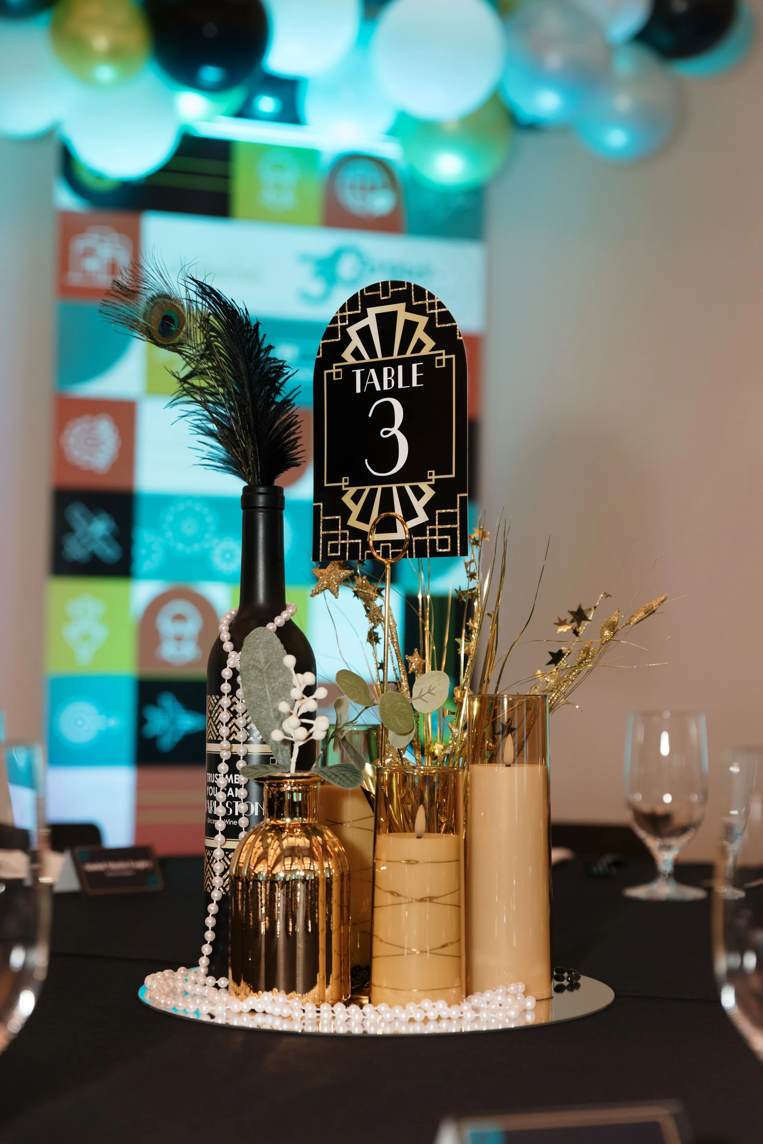 Table centerpiece with gold and black vases, peacock feather, gold star decorations, and a sign that says 'Table 3', set on a black table with glassware and a mirrored base.