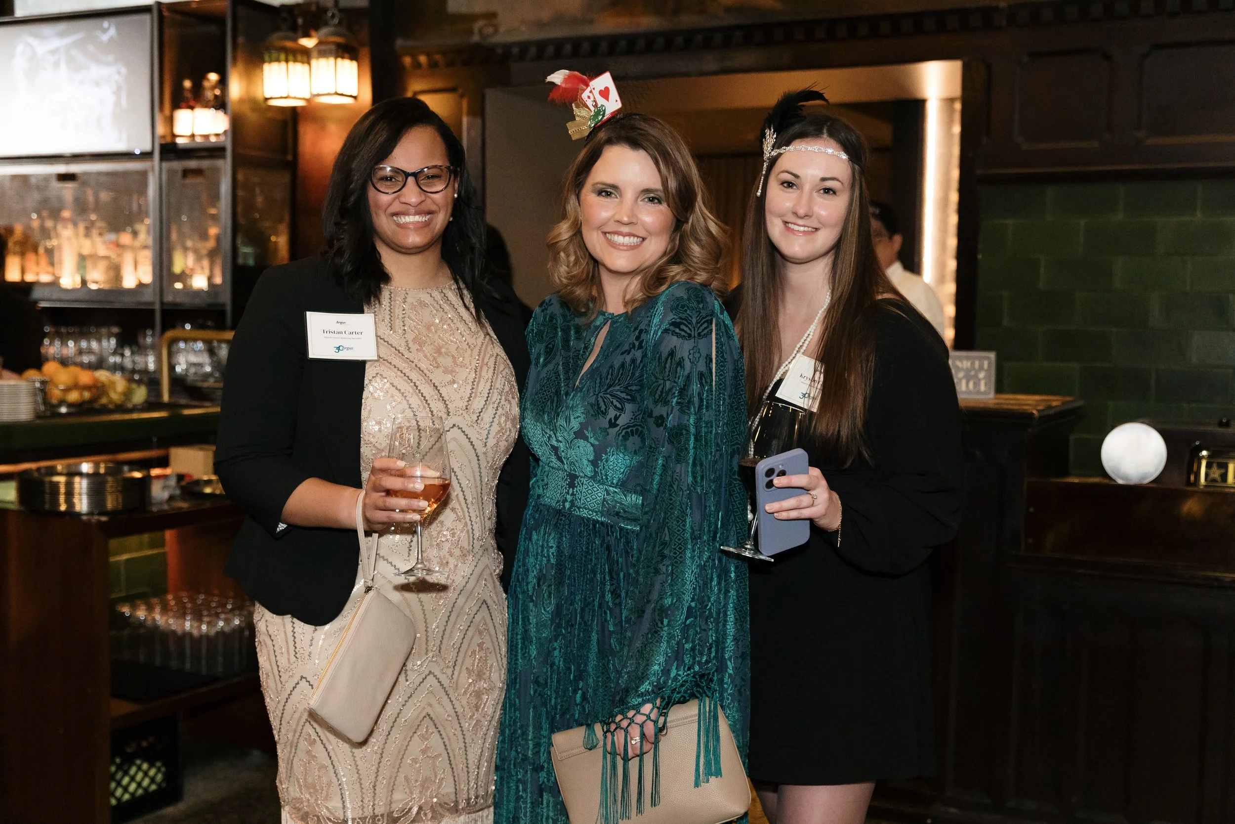 Three women in formal attire at an indoor social event, one holding a wine glass and wearing a name tag, standing close together and smiling at the camera, with a bar and warm lighting in the background.
