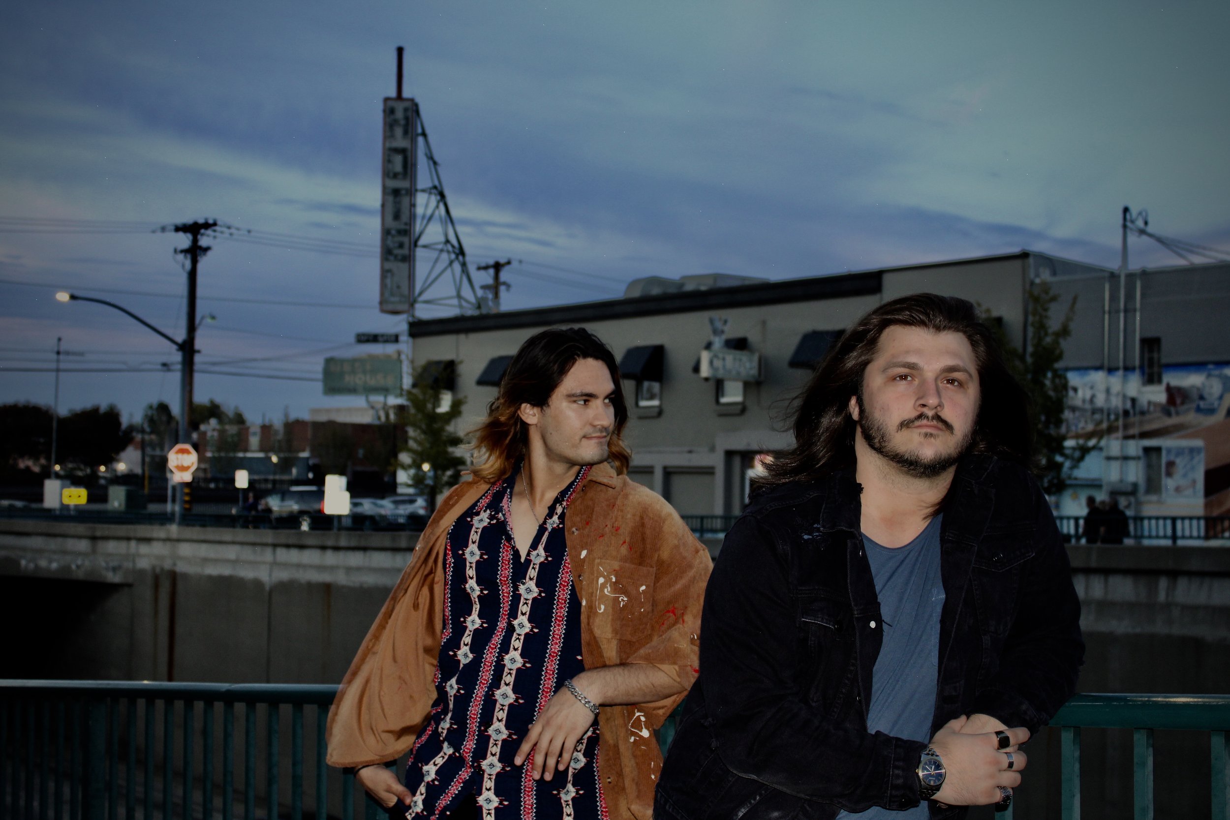 Two men standing on a bridge with an urban backdrop at dusk.