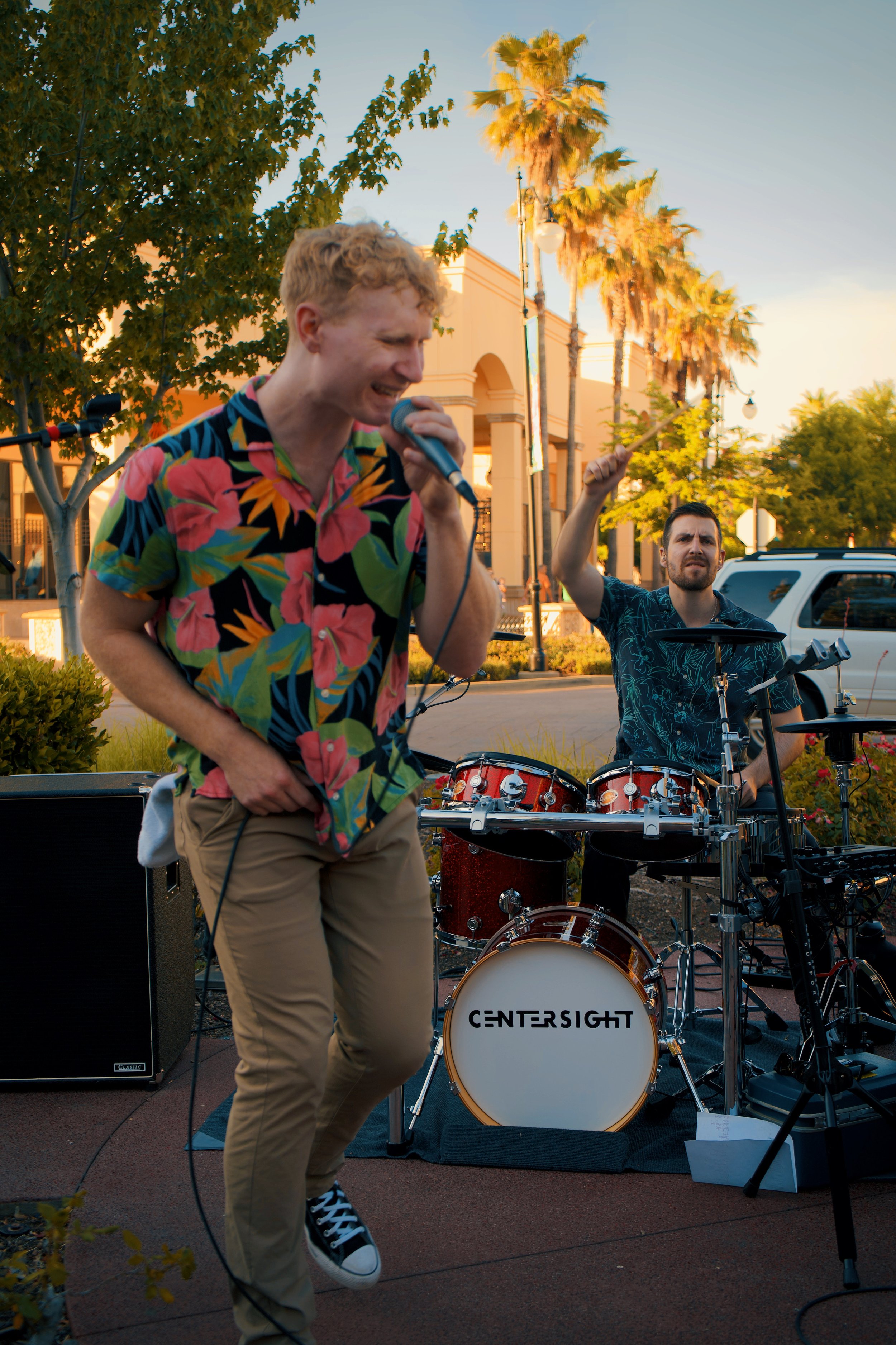 Outdoor band performance with a singer in a floral shirt holding a microphone and a drummer playing a drum set. Trees and buildings in the background.