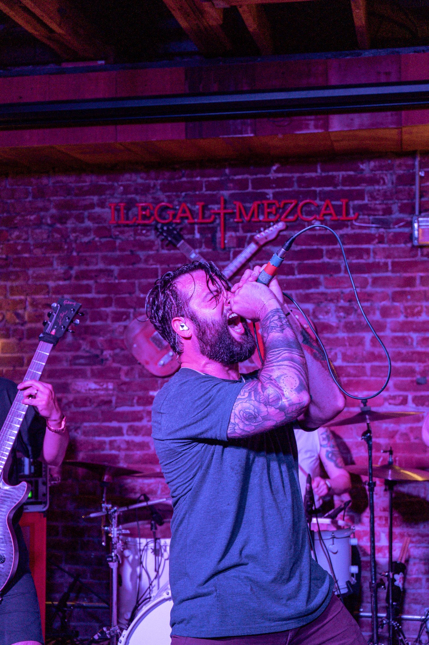 Musician performing passionately on stage with microphone, tattooed arms, electric guitar, and drums in background, under Ilegal Mezcal sign.