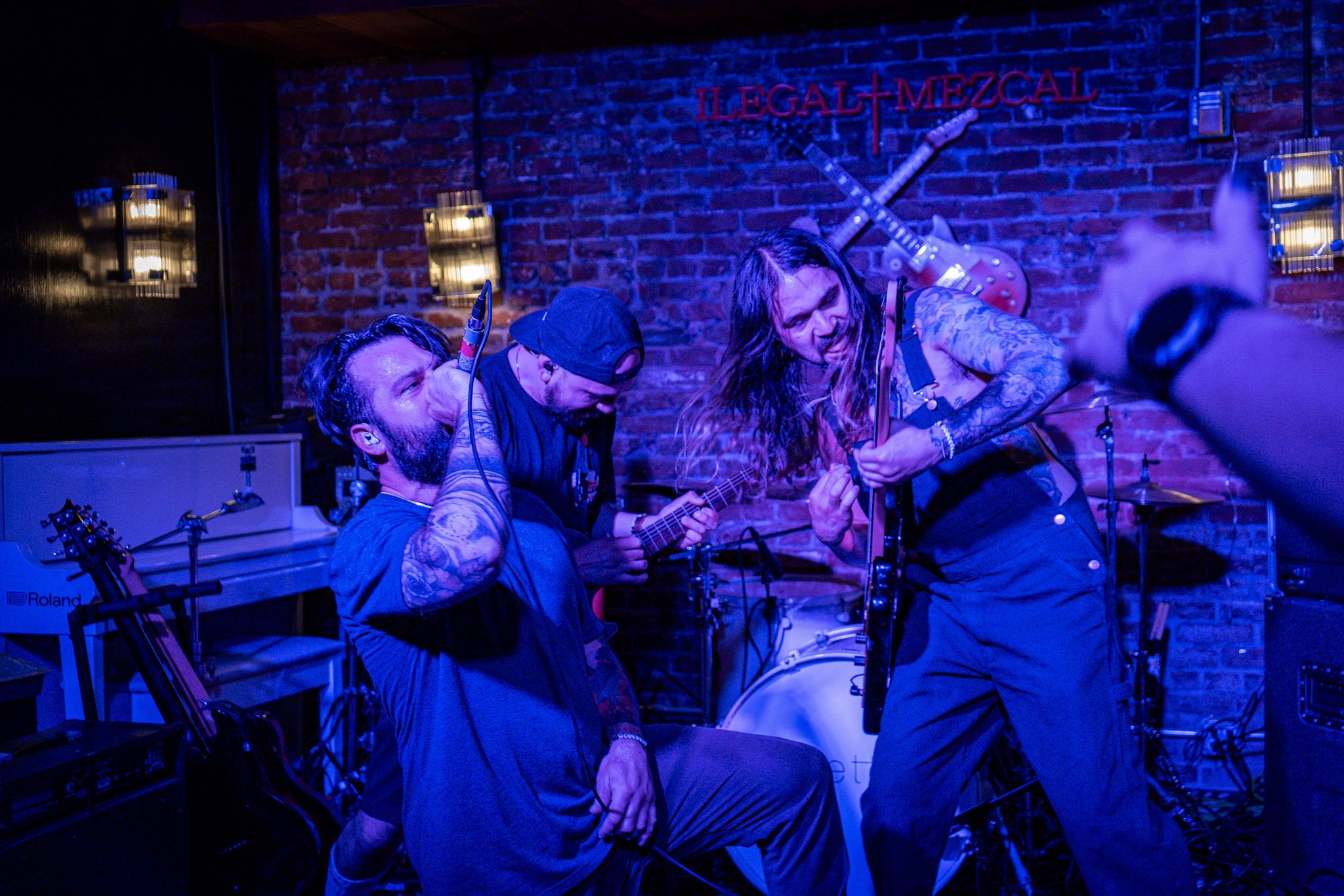 A live rock band performance in a small venue with musicians playing guitar and singing passionately. The background features a brick wall and a drum set, with guitars hanging as decor. The lighting is vibrant and blue.