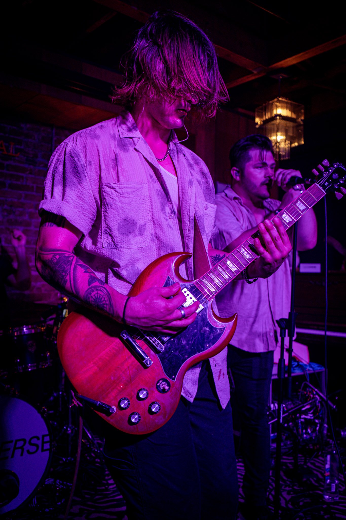 Musician playing electric guitar on stage with another person singing into a microphone, colored stage lights, and musical equipment visible.