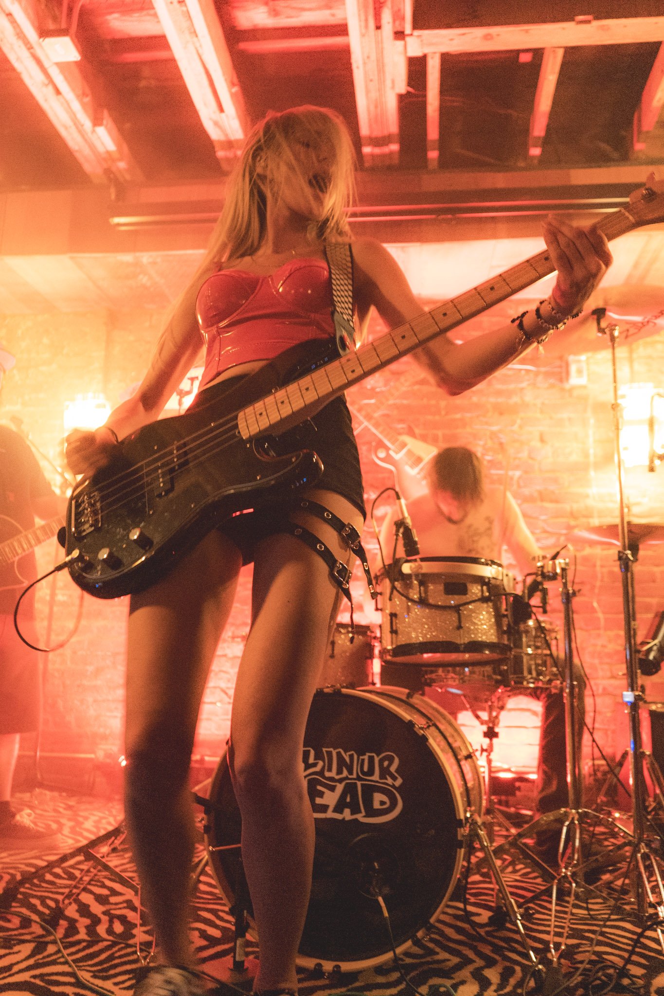 A female musician playing a bass guitar on stage with a drummer in the background, under warm stage lighting.