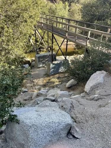 A wooden bridge crossing over a small creek in a wooded area, with rocks and trees surrounding it.