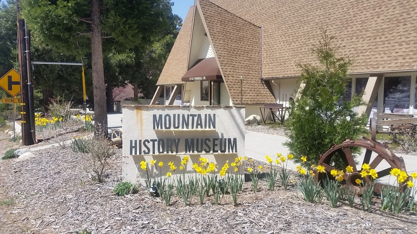 Sign reading 'Mountain History Museum' in front of a landscaped area with yellow flowers, a small tree, and an aged wagon wheel, near a building with a sharply angled roof.