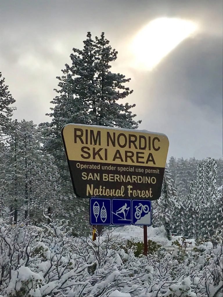 Snow-covered trees and bushes under a cloudy sky with the sun partially visible behind clouds. A sign indicating the Rim Nordic Ski Area in San Bernardino National Forest, with icons for snowshoeing, cross-country skiing, and mountain biking.