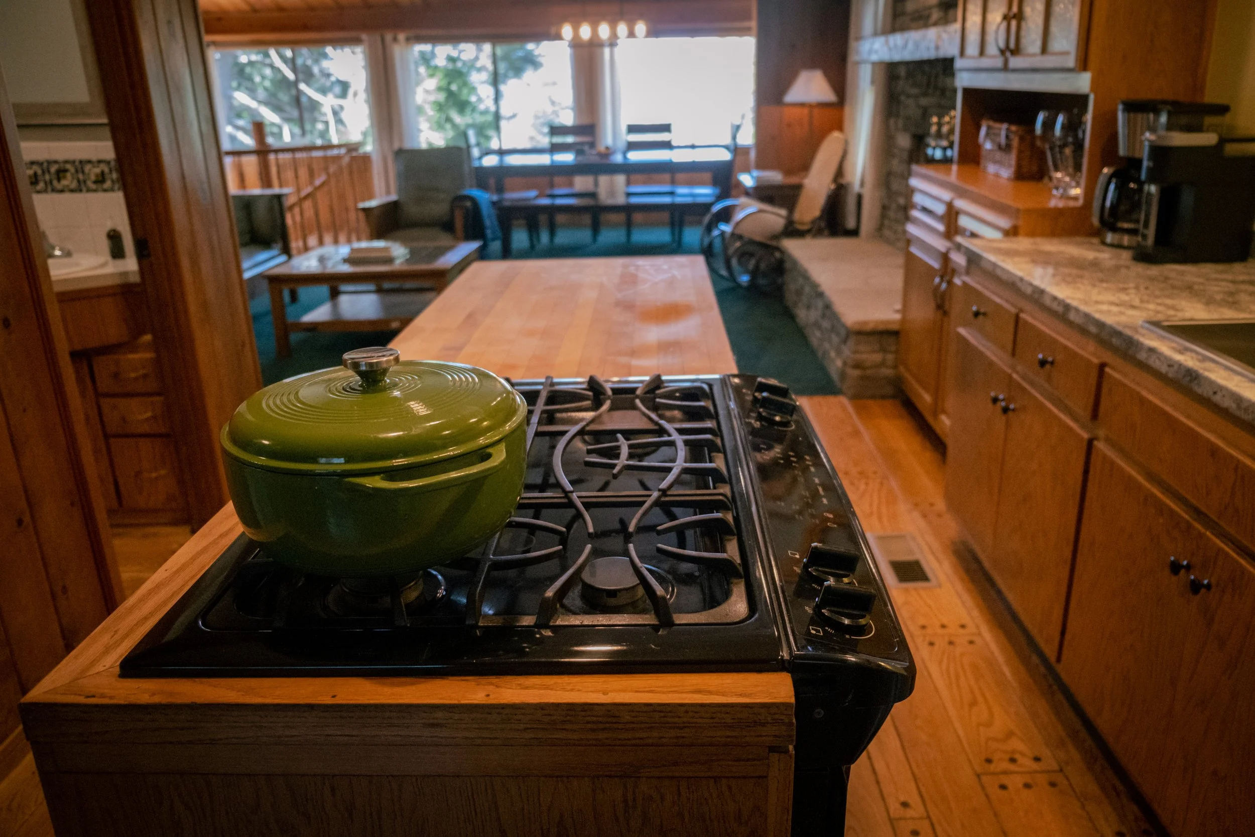 View from kitchen stove showing a green pot on the stove, with a dining and living area in the background featuring large windows and wooden furnishings.