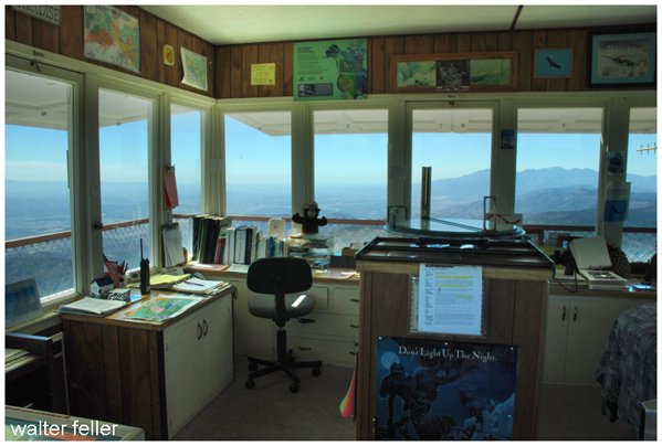 Observation room with large windows overlooking mountains, with desks, books, a computer, and a chair.