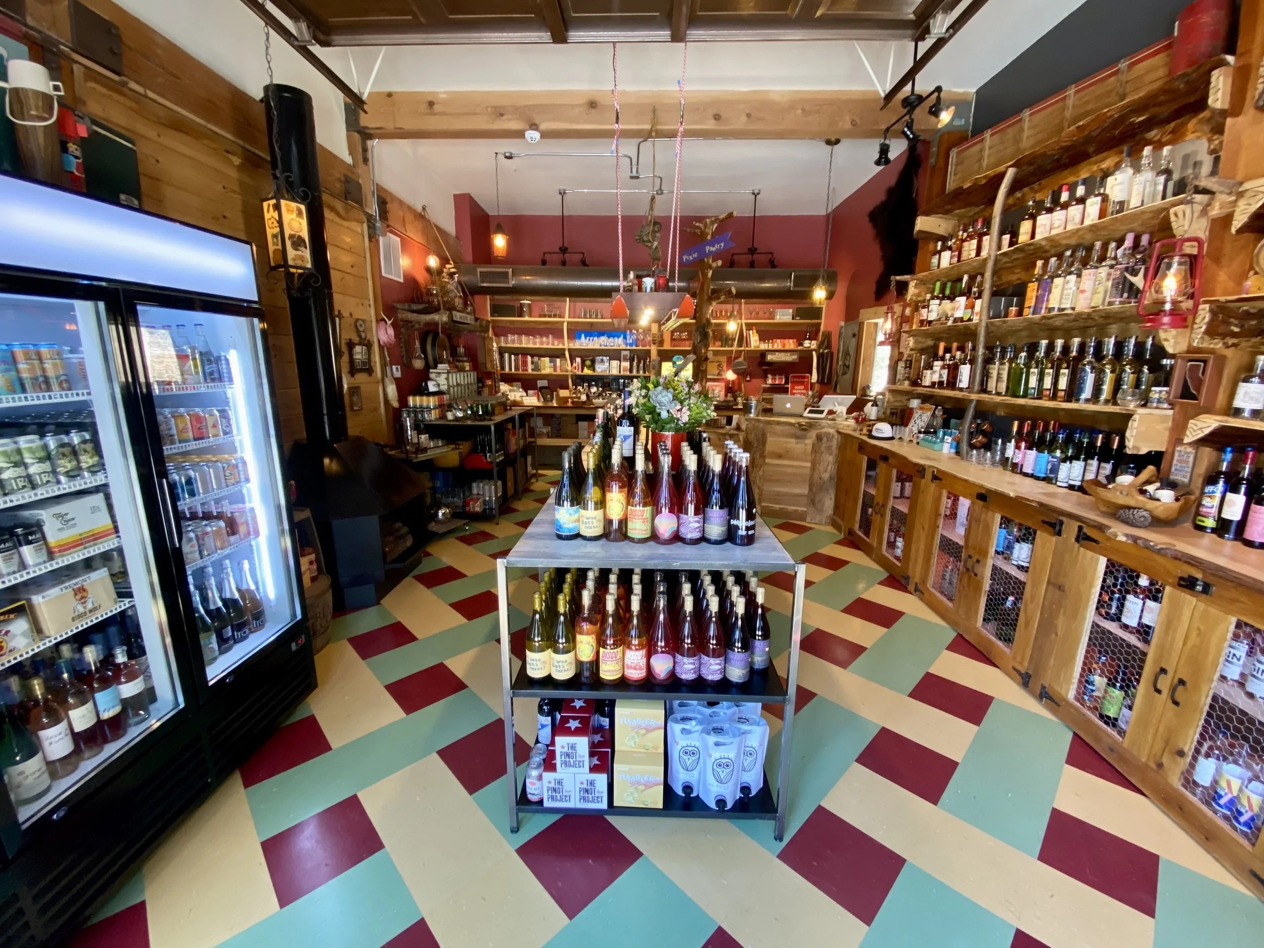 Interior of a cozy shop or café with wooden walls and shelves, bottles of beverages on a central display, a fridge filled with drinks on the left, and decorative elements like flowers and artwork on the walls.