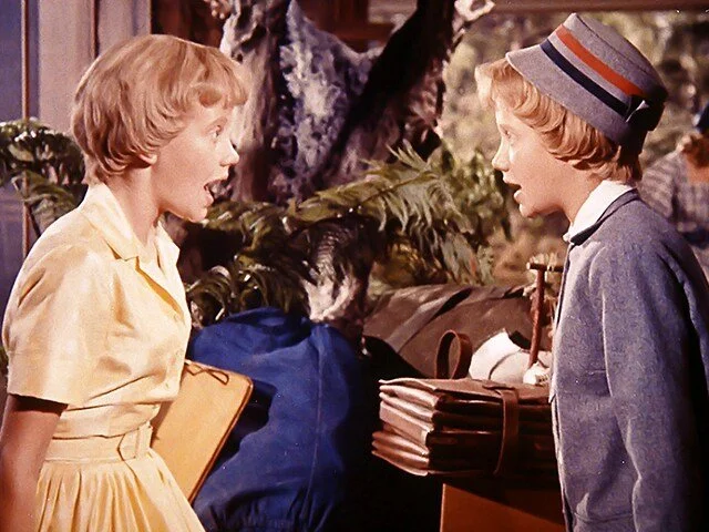 Two children, a girl in a yellow dress and a boy in a blue jacket and gray hat, appear to be talking to each other indoors, possibly at a school or a camp, with trees and other people in the background.