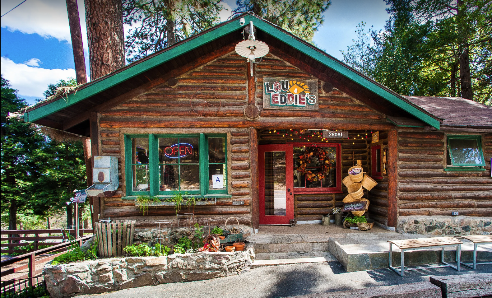 A rustic log cabin with a green roof, colorful string lights, decorative wreath, and plants in front. The house has signs including an 'Open' neon sign in the window, a sign reading 'Lou Eddies,' and an address number 28561. The scene is sunny with trees and a blue sky in the background.