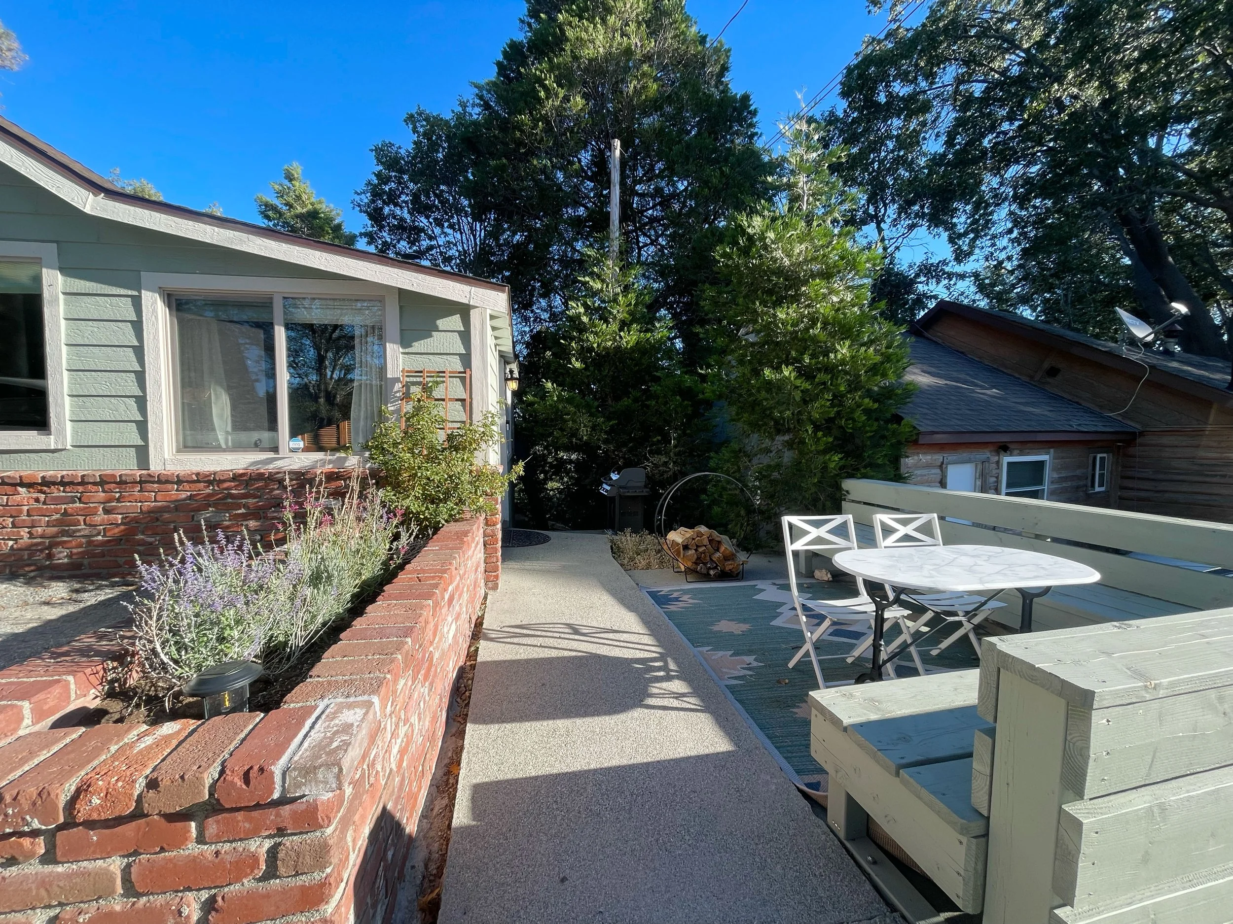 A backyard patio featuring a brick planter with lavender plants, a white wooden table with chairs, and houses surrounded by trees under a blue sky.