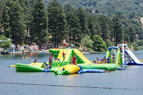 People enjoying an inflatable water park on a lake with covered picnic areas and trees in the background.