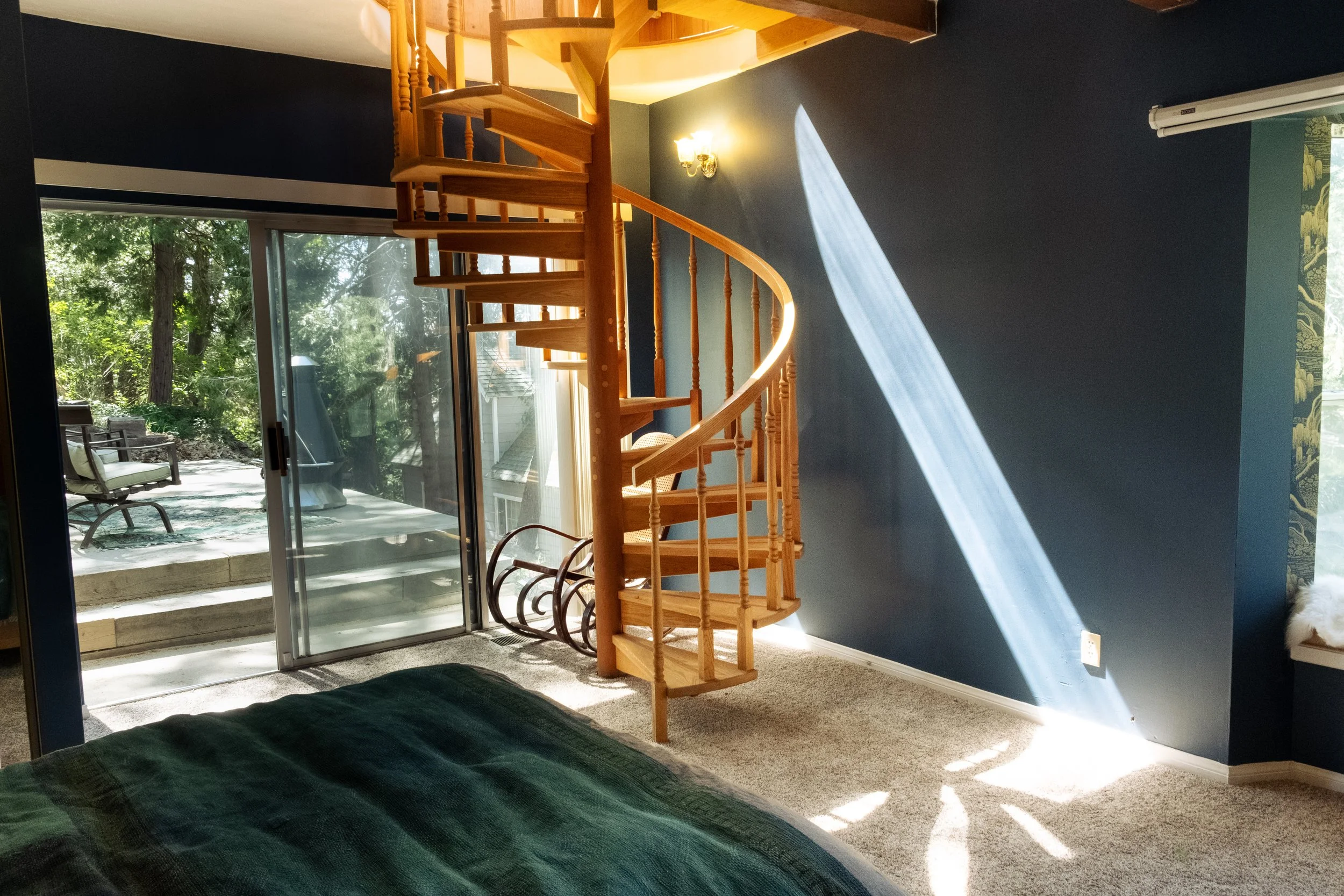 Living room with a wooden spiral staircase, large sliding glass door leading to an outdoor patio with chairs, and a wall-mounted light fixture. Sunlight streams through the window creating a bright and airy atmosphere.