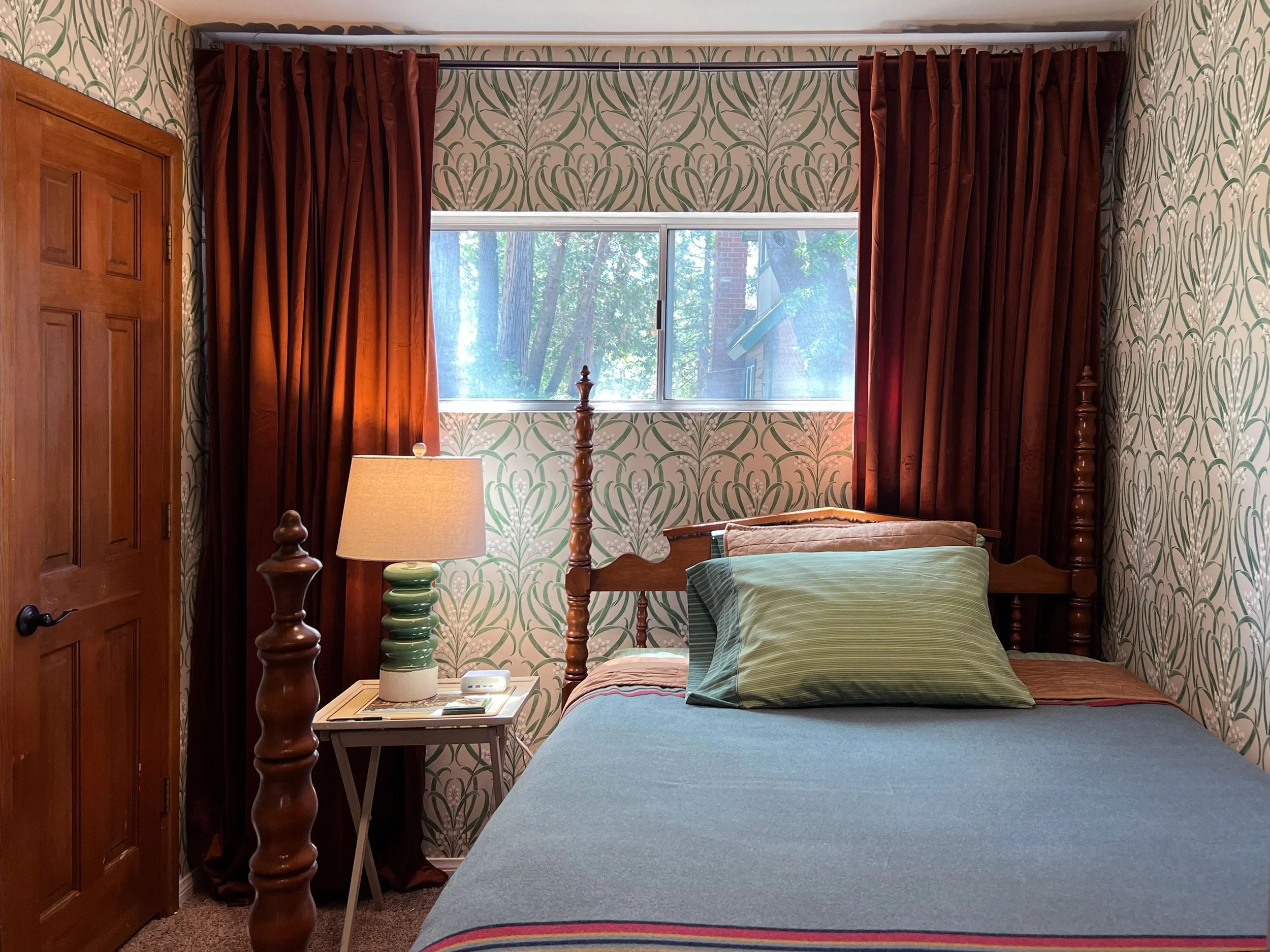 A bedroom with a wooden bed, green-striped bedding, a bedside table with a lamp, a window with floral curtains, and patterned wallpaper.