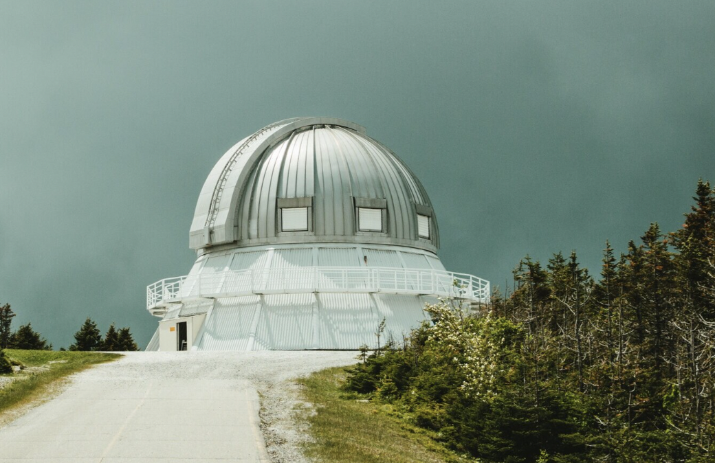 A large observatory with a metallic dome, situated on a hilltop with a gravel road leading up to it, surrounded by trees under a cloudy sky.