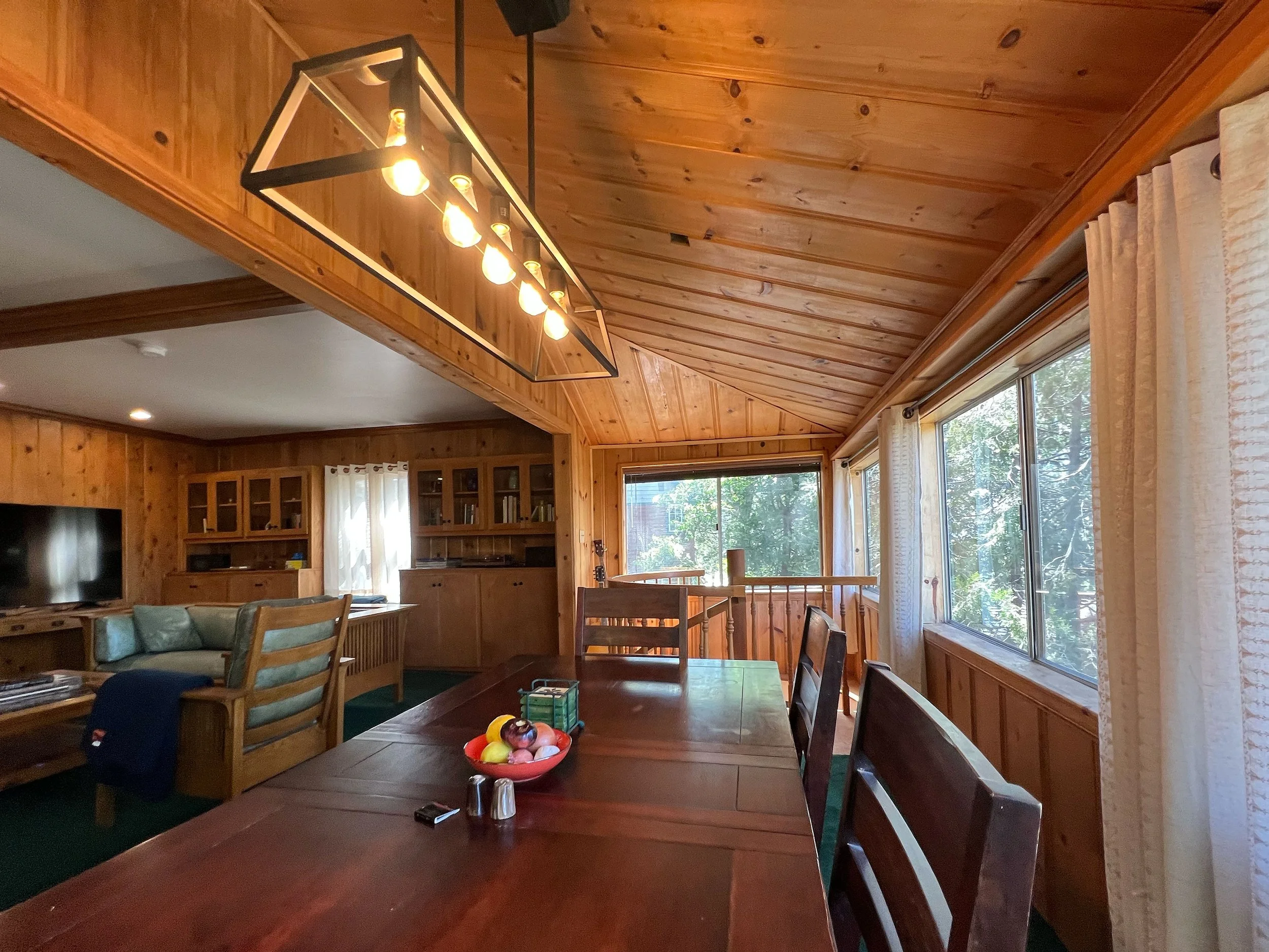 A dining room with wooden walls, ceiling, and furniture. A large window with curtains on the right side, allowing natural light. A chandelier with multiple bulbs hanging from the ceiling. A dining table with chairs, and a fruit bowl in the center. In the background, a living area with a television and cabinets.