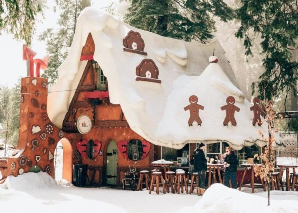 A festive gingerbread house decorated with snow and gingerbread figurines, with trees and snow in the background and people standing nearby.