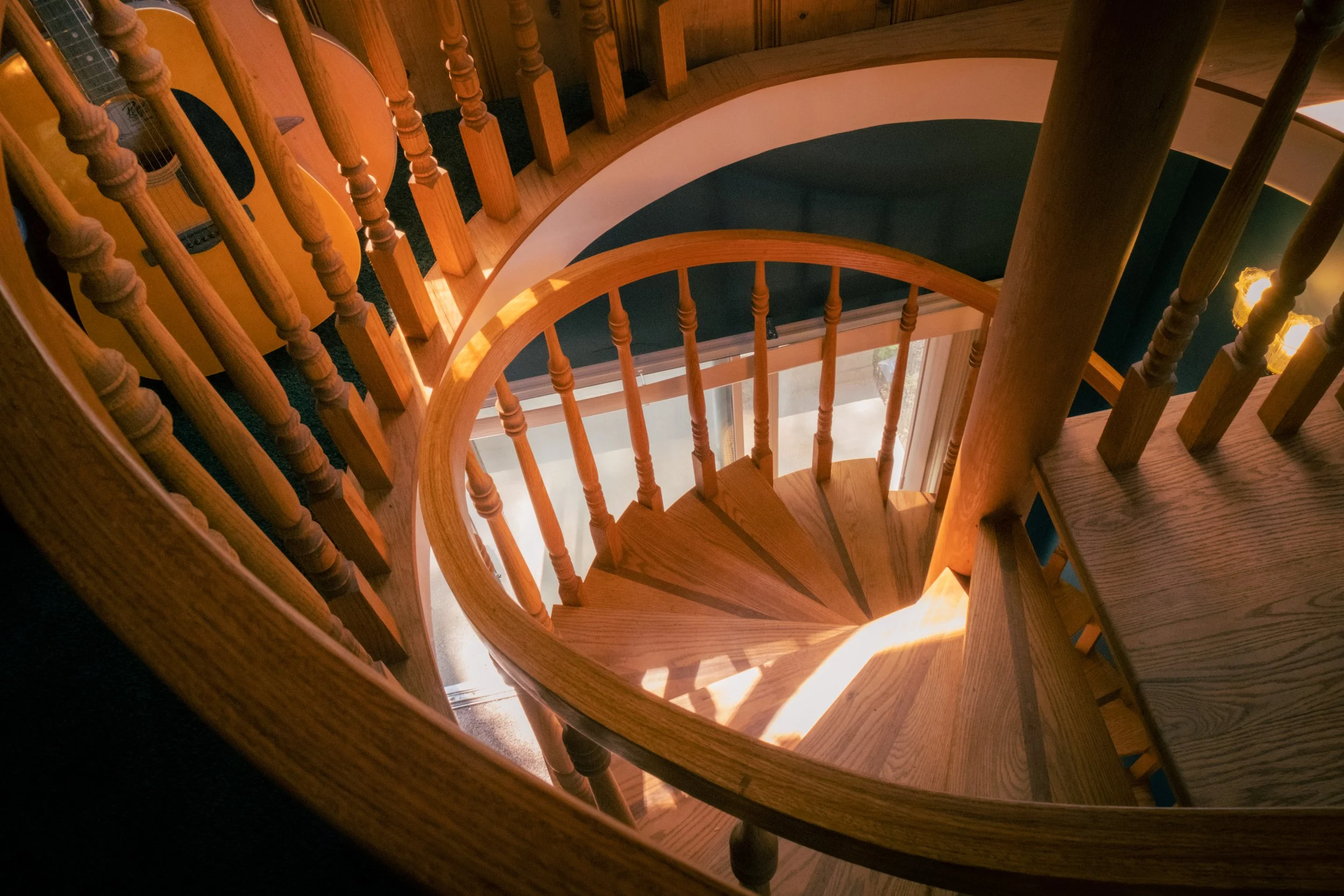 Spiral staircase made of wood with a black carpeted step in the middle, viewed from above, with natural light coming through a window.