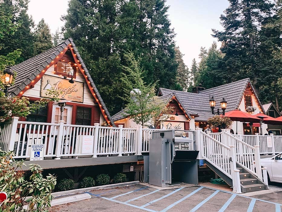A cozy restaurant with a chalet-style exterior, surrounded by trees, with string lights and outdoor seating, and a white picket fence.