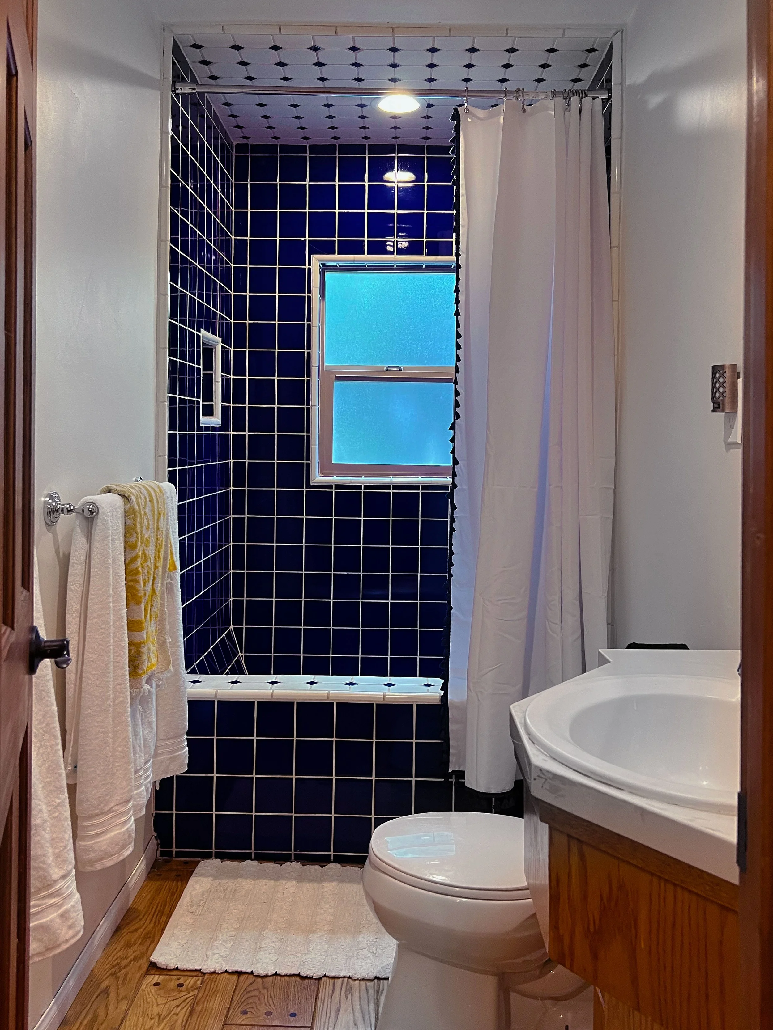 Bathroom with black tiled shower area, frosted window, white toilet, wooden vanity with sink, beige towels, and white curtains.