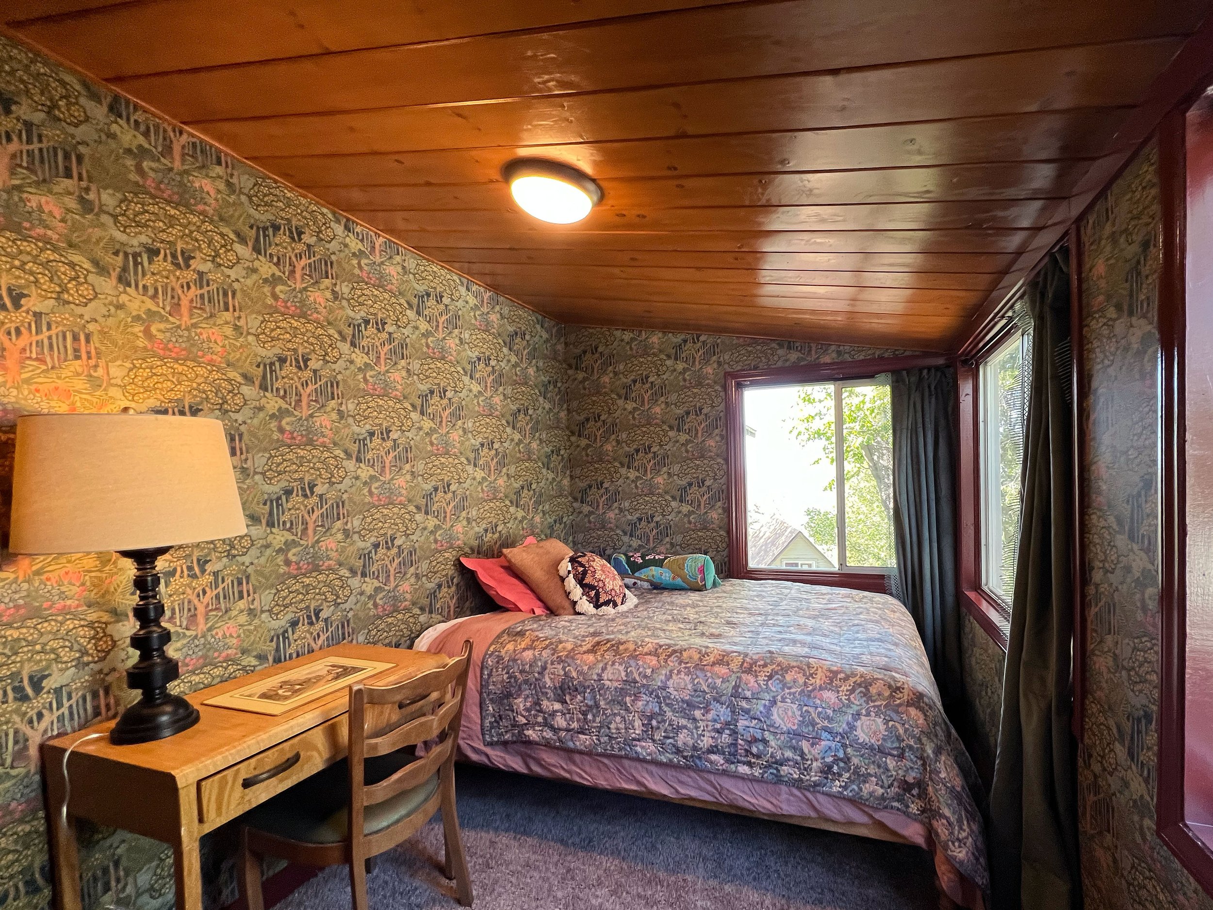 Interior of a bedroom with a wood ceiling, floral wallpaper, a bed with multiple pillows, a window with curtains, a bedside table with a lamp, and a wooden chair.