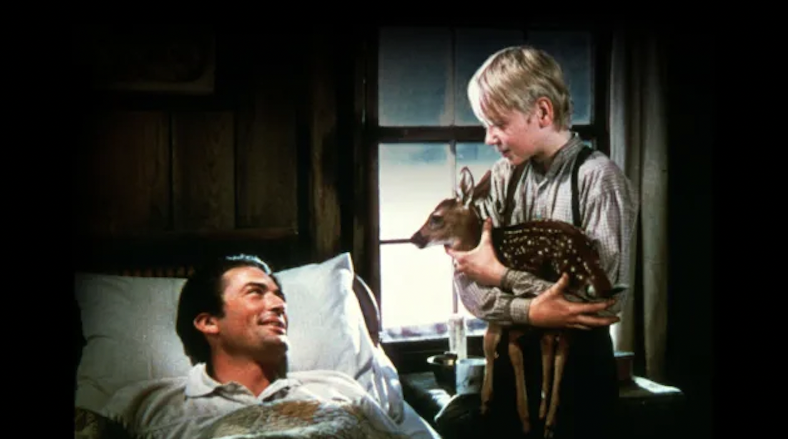 A man lying in bed with an amused expression looking at a young boy standing next to him, holding an opossum, inside a cozy room with a window and wooden walls.