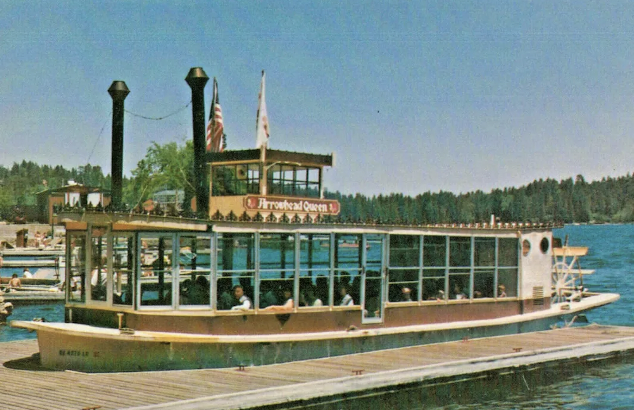 A boat with passengers on a lake or river, with a wooden dock in the foreground. The boat has a glass-enclosed upper deck, a sign that reads 'Freshette Queen', and two American flags. Tall trees and a hill are visible in the background under a clear blue sky.