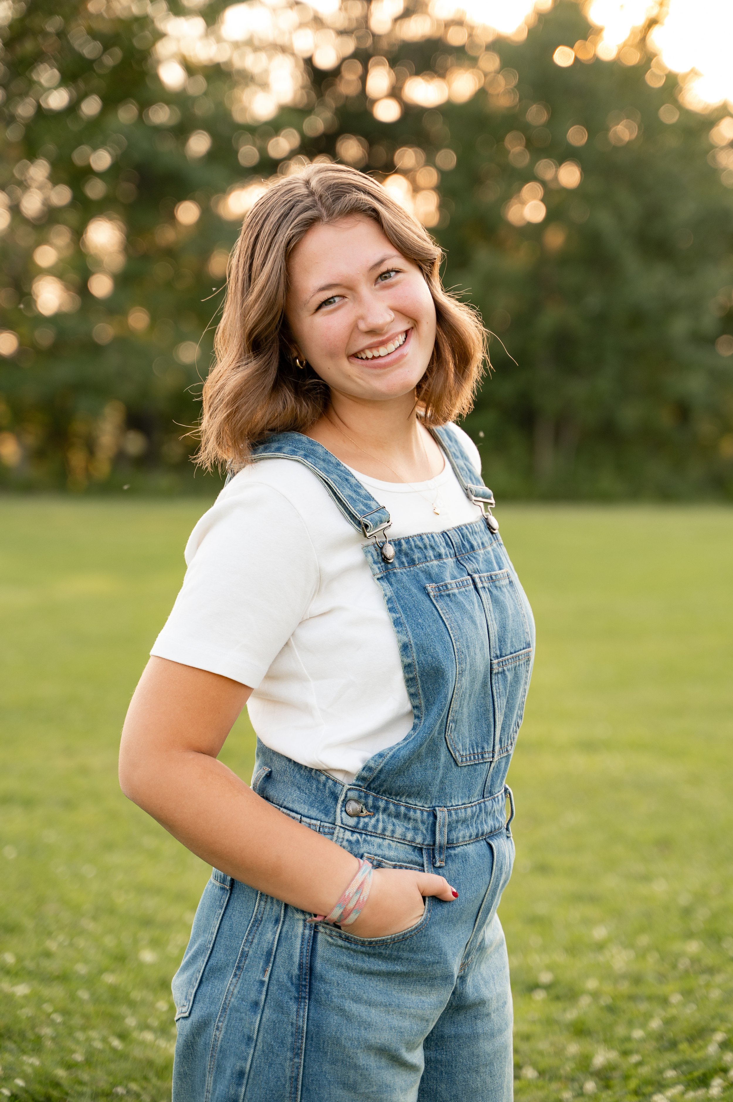 A young woman smiling outdoors during sunset, wearing a white t-shirt and denim overalls.