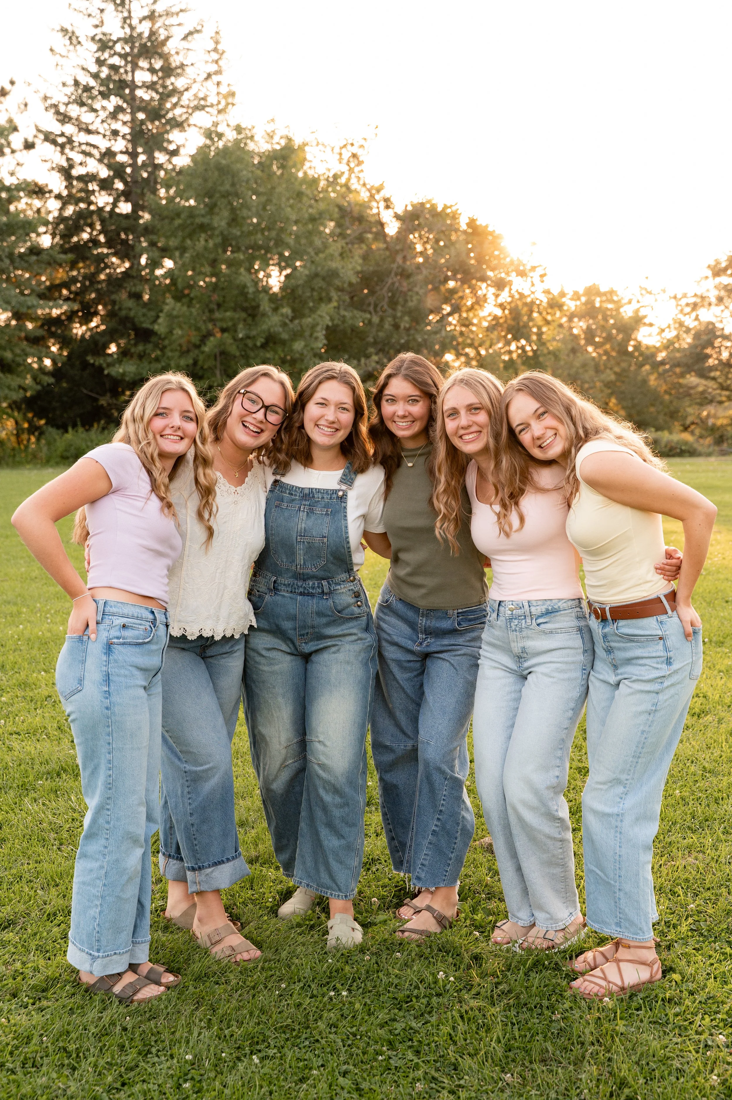 A group of six young women standing close together on grass during sunset, smiling, with trees in the background.