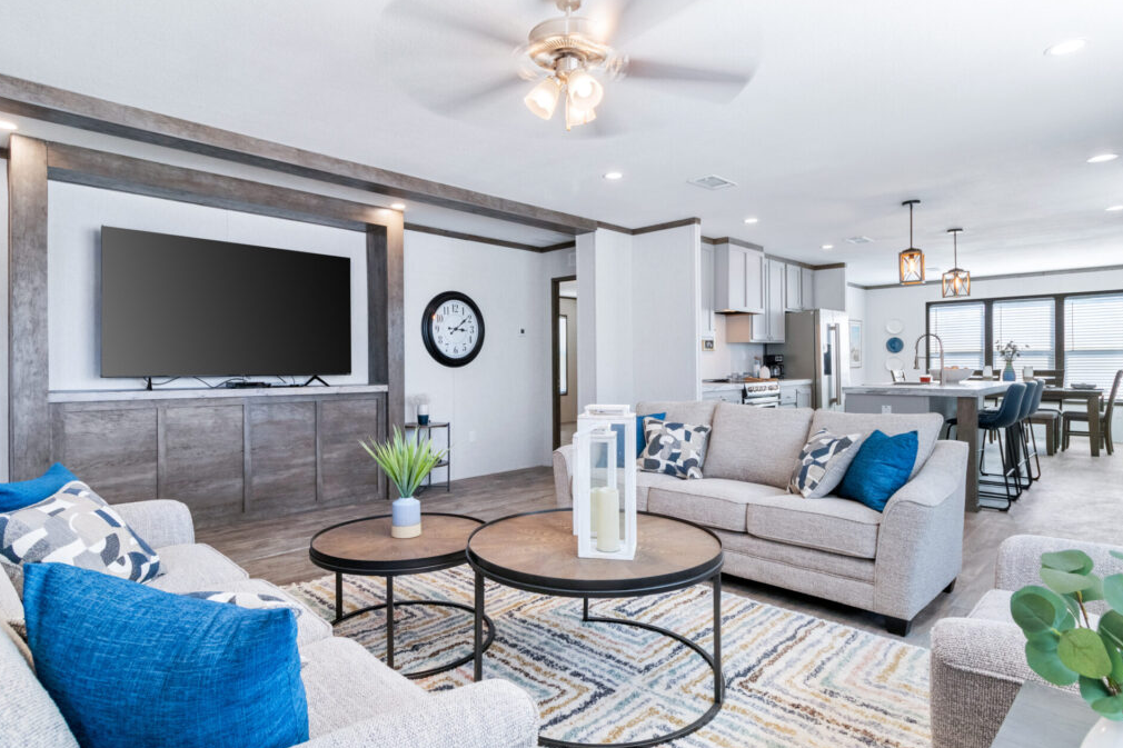 Open-concept living room and kitchen with beige sofas, blue and patterned pillows, wooden coffee tables, a wall-mounted TV, a round clock, decorative lanterns, and a dining area with barstools.
