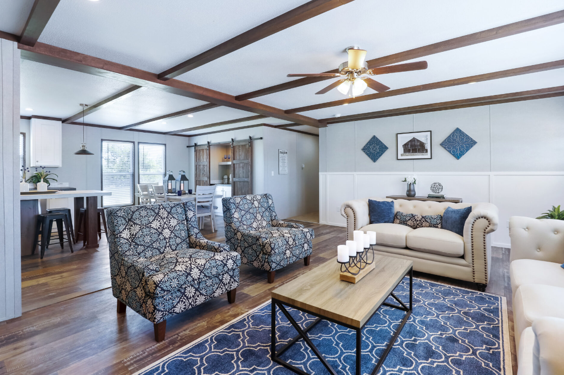 Living room with white walls, wooden ceiling beams, a ceiling fan, a white couch with blue and patterned pillows, two patterned armchairs, a wooden coffee table with candles, a blue patterned rug, and a dining area with a table and chairs in the back
