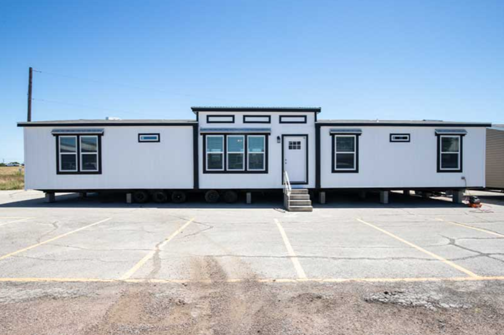 White tiny house on wheels with black window frames, set in a parking lot under a clear blue sky.