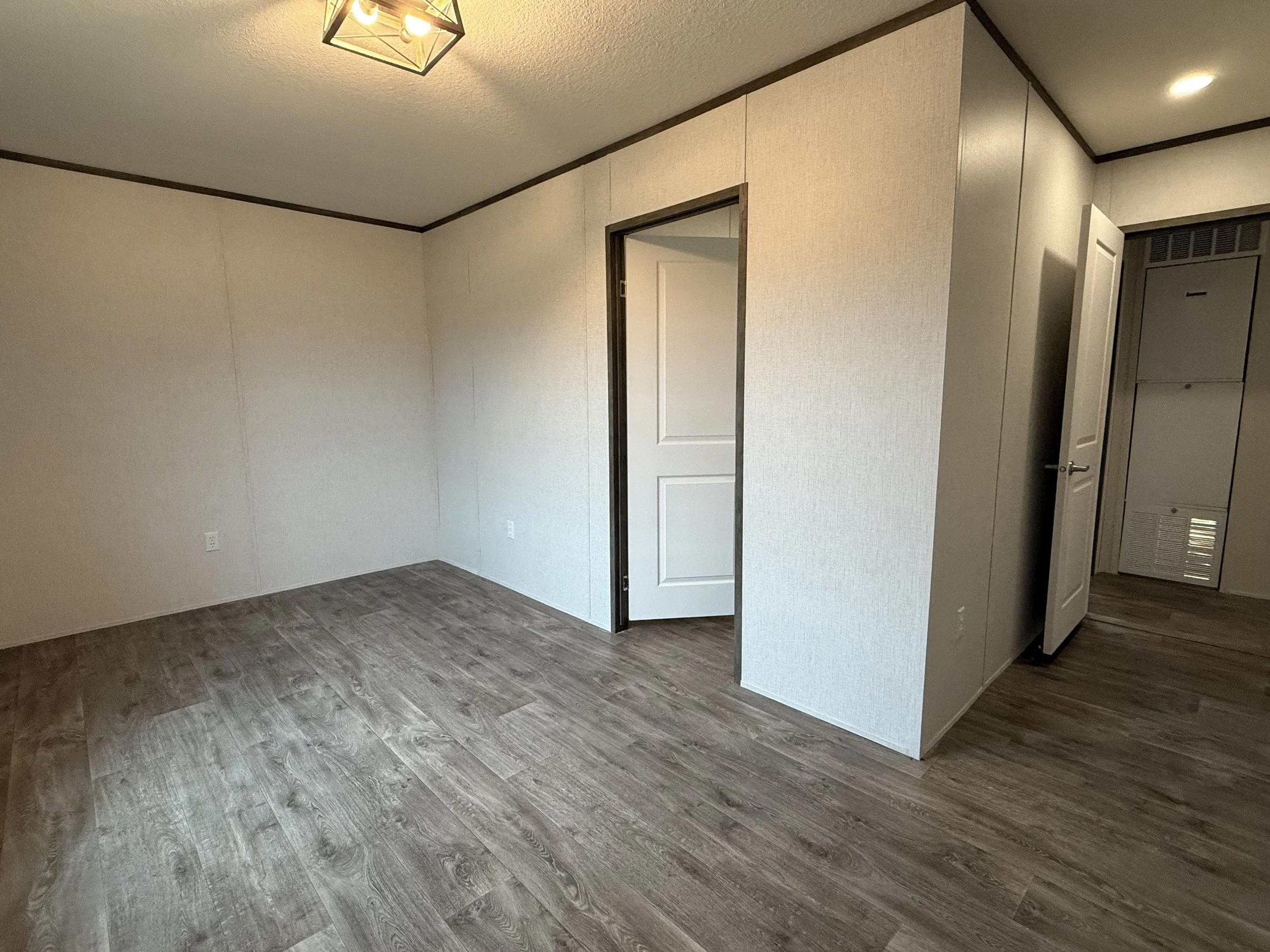 Empty room with wood laminate flooring, white textured walls, a ceiling light fixture, and a door leading to another room.