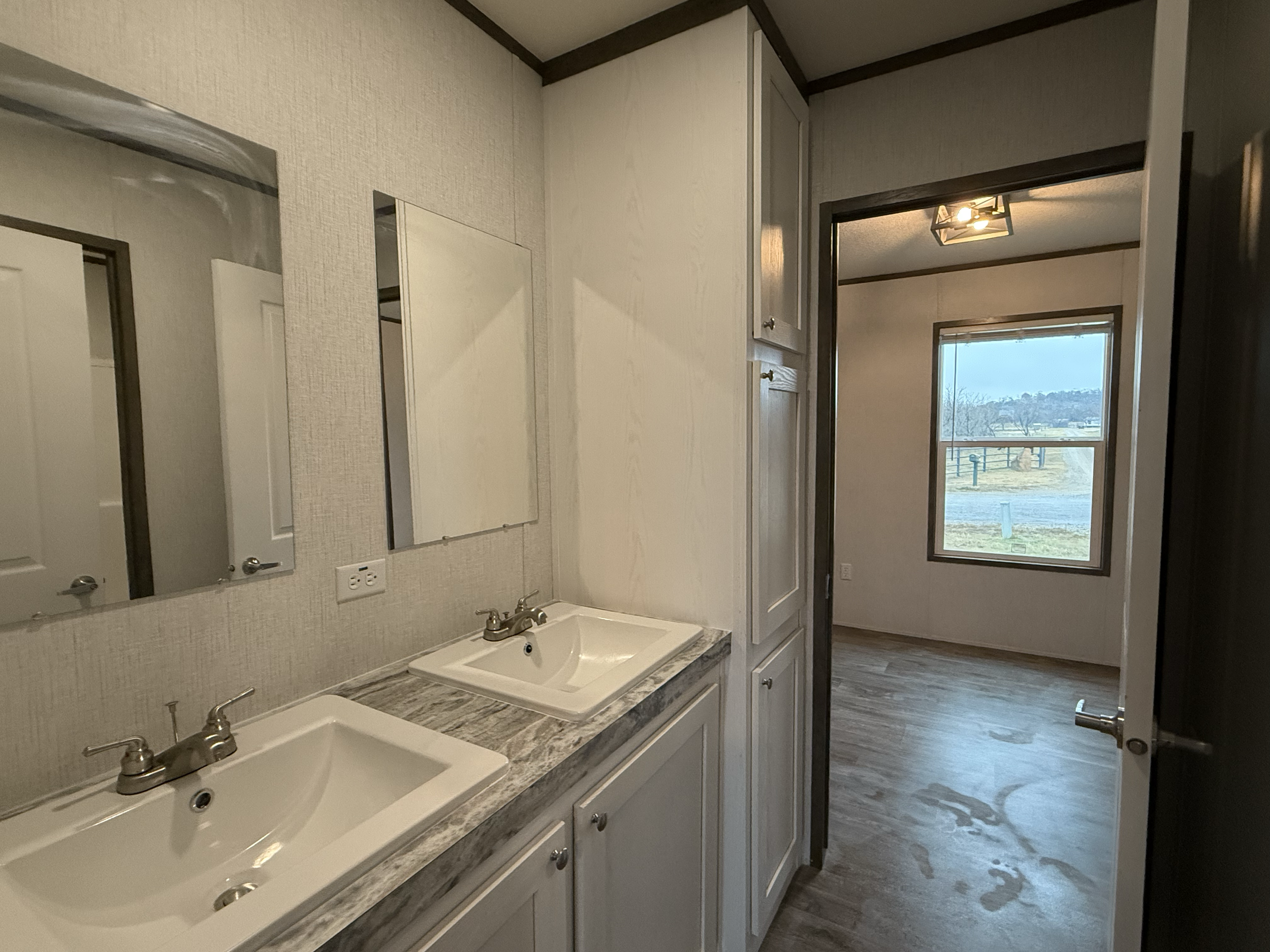 Bathroom vanity with two sinks, mirrors above, as well as a closet and a doorway leading to a room with a window and wood-like flooring.
