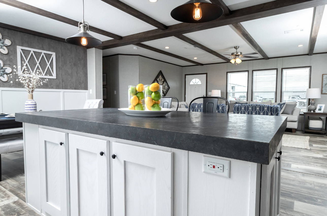 Modern kitchen with a black countertop island, decorative fruit centerpiece, and a living area with large windows, sofas, and lamps.