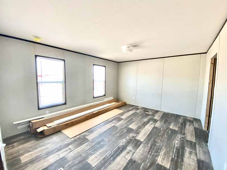 Empty room with light gray walls, two windows with blinds, a ceiling light, and a dark wood floor. Construction materials are stacked against the wall.