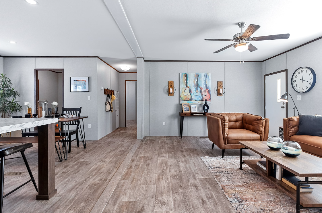 Living room with wood flooring, a tan leather sofa, a wooden coffee table with decorative items, wall art featuring guitars, a clock, a ceiling fan, and a doorway leading to other rooms.